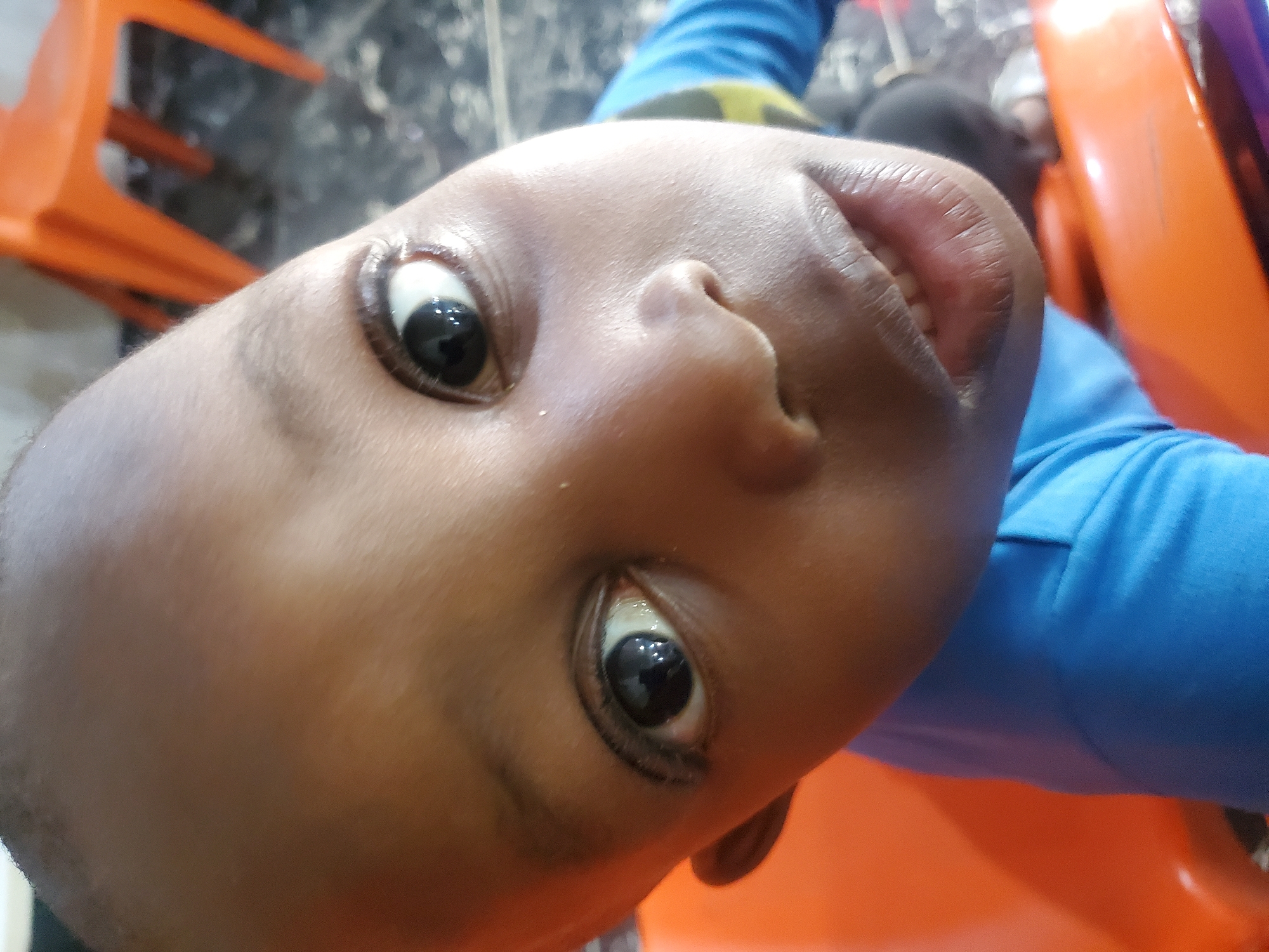 A close-up, high-angle shot shows the face of a young Black child. The child is looking directly into the camera with wide, dark eyes and a slight smile. The child's head is shaved. The child is wearing a blue shirt with a yellow and black stripe across the chest. The background is out of focus but appears to be an indoor setting with an orange chair visible. The overall lighting is bright. The image is a photograph taken with a mobile device.