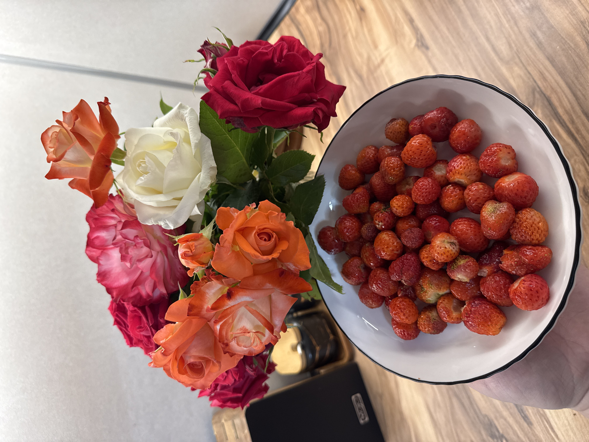 An overhead shot shows a hand holding a white bowl filled with strawberries. The bowl has a black rim. Behind the bowl, a bouquet of roses in shades of red, pink, orange, and white is arranged in a vase. The background consists of a wooden countertop and a blurred wall.