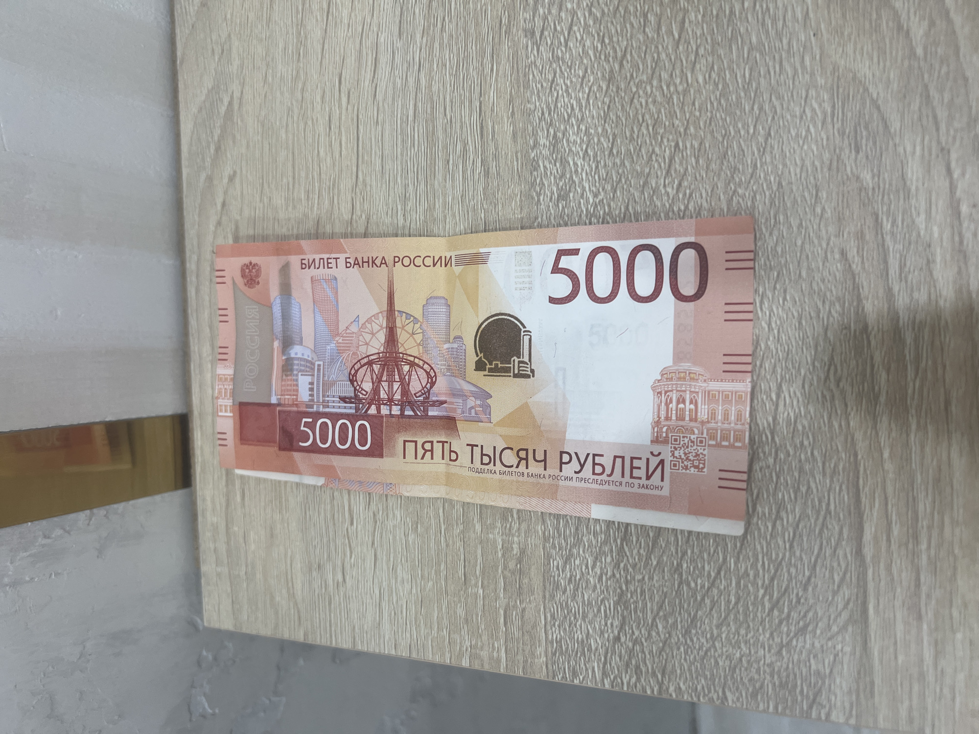 A Russian 5000 ruble banknote is lying on a wooden surface. The banknote is oriented vertically with the top edge towards the right. The denomination "5000" is prominently displayed in large red numerals on the left side of the banknote, and also in red on the right side. Text in Russian, including "БИЛЕТ БАНКА РОССИИ" (Bank of Russia Ticket) and "ПЯТЬ ТЫСЯЧ РУБЛЕЙ" (Five Thousand Rubles), is visible. The background consists of a light gray textured wall and a light-colored wooden surface, possibly a table or shelf. There are no people or animals in the image. The lighting appears to be natural or from an overhead source, casting subtle shadows. The scene is static and appears to be an indoor environment. There is no information within the image that can identify the specific location as "Veteran-2, Russia."