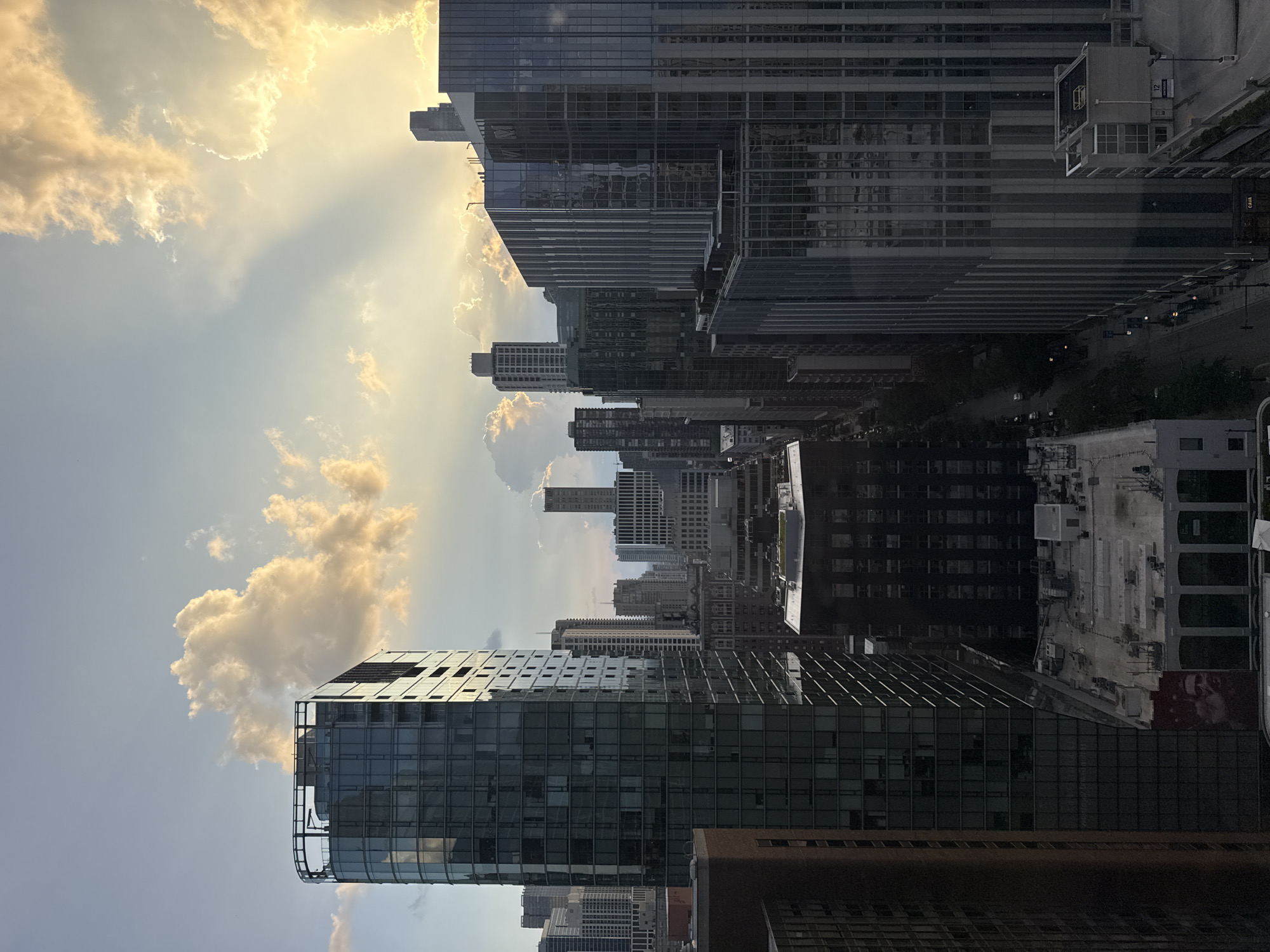 This is a vertical, eye-level, wide-angle shot of a cityscape in Chicago, Illinois, USA, during a sunset. The sky is filled with dramatic clouds, some of which are illuminated by the setting sun, casting rays of light downwards. The dominant feature of the image is a cluster of modern skyscrapers, characterized by their glass facades and sharp architectural lines.
In the foreground, on the left, is a tall, slender building with a dark, reflective glass exterior and a distinct curved top. Adjacent to it, to the right, is another large skyscraper with vertical ribbed detailing on its glass facade, reflecting the sky and surrounding buildings. Further into the cityscape, a dense arrangement of various other skyscrapers, some taller and some shorter, create a layered urban panorama. Some of these buildings have a more traditional architectural style with visible windows, while others continue the modern glass aesthetic.
The street level is visible in some areas, showing a road with sparse traffic and trees lining the sidewalks. The overall impression is one of a bustling metropolis viewed from a high vantage point, with the natural beauty of the sunset adding a dynamic element to the urban landscape. The lighting suggests it is either early morning or late evening.