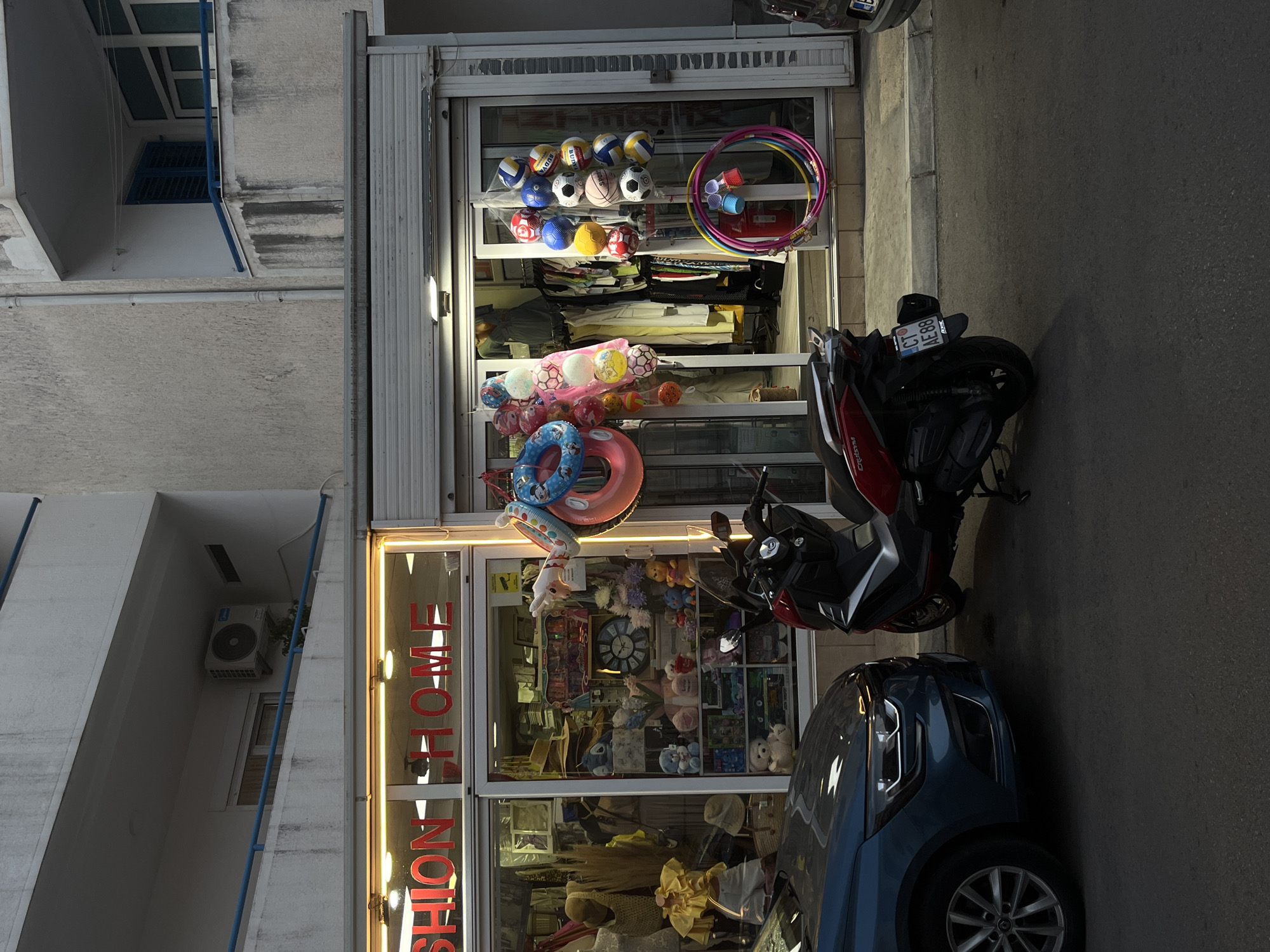 The image depicts a street scene in Budva, Montenegro, with a focus on the storefront of a shop. The shop's signage, illuminated by warm-toned lights, reads "FASHION HOME". The display windows are filled with various items, including clothing on mannequins, stuffed animals, toys, and decorative items. To the right of the shop entrance, a cluster of colorful inflatable rings and balls are displayed outside. A red and black scooter is parked in front of the shop, and a blue car is partially visible on the left. The building appears to be multi-story, with balconies and an air conditioning unit visible on the upper levels. The street itself is paved and appears to be a pedestrian or low-traffic area. The overall lighting suggests it is either dusk or early morning.