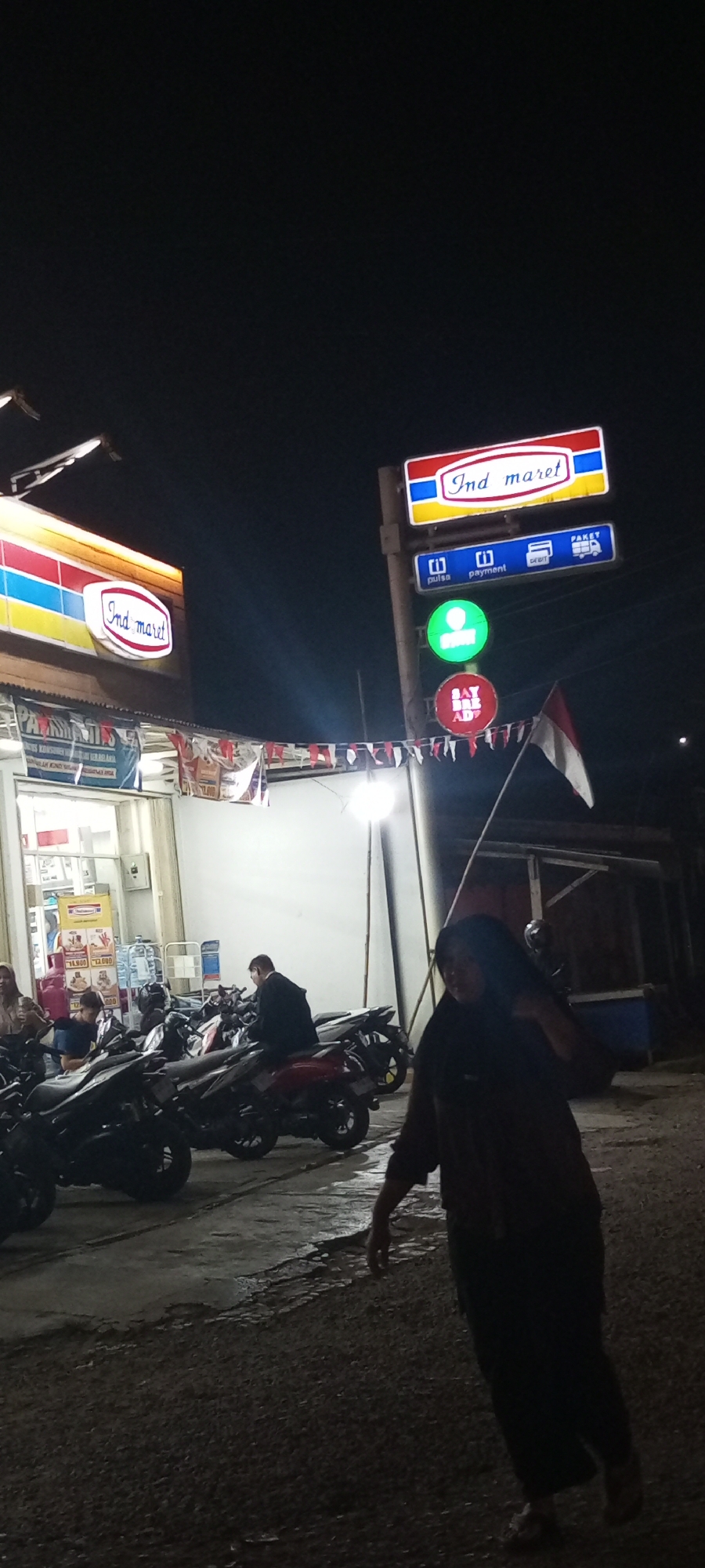 The image depicts a nighttime scene outside an Indomaret convenience store in Setu, Indonesia. A large, illuminated Indomaret sign is visible at the top of a pole, accompanied by smaller signs indicating "pulsa," "payment," "DEBIT," and "PAKET." Below these, a green circular light and a red circular light are present. A person wearing a dark hijab and long clothing walks in the foreground. In the background, several motorcycles are parked in front of the store. A man in a black jacket is seated on one of the motorcycles. The store's entrance is lit, and merchandise is visible inside. Red and white Indonesian flags are strung across the front of the store. The ground appears wet in some areas, suggesting recent rain.