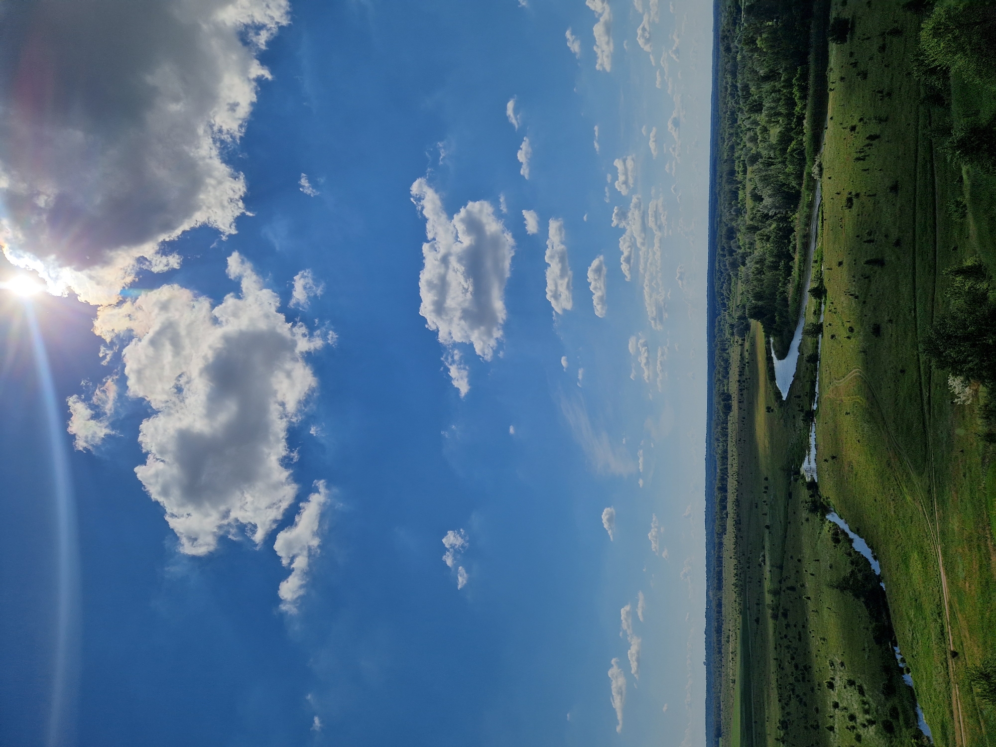 The image depicts a wide-angle landscape under a bright, sunny sky with scattered clouds. The sun is visible in the upper right corner, casting rays and creating a lens flare effect. The scene is a rural panorama with rolling green fields, a winding river, and a distant treeline. There are no people or man-made structures immediately visible in the foreground or mid-ground, suggesting a natural environment. The terrain is characterized by lush vegetation, with variations in shade and texture across the fields. A narrow, dark blue river meanders through the landscape, reflecting the sky. The horizon is marked by a dense forest, beyond which lies a hazy expanse under the bright sky. The overall impression is one of a serene and expansive natural landscape on a clear day. The location is identified as Ustyvytsia, Ukraine.