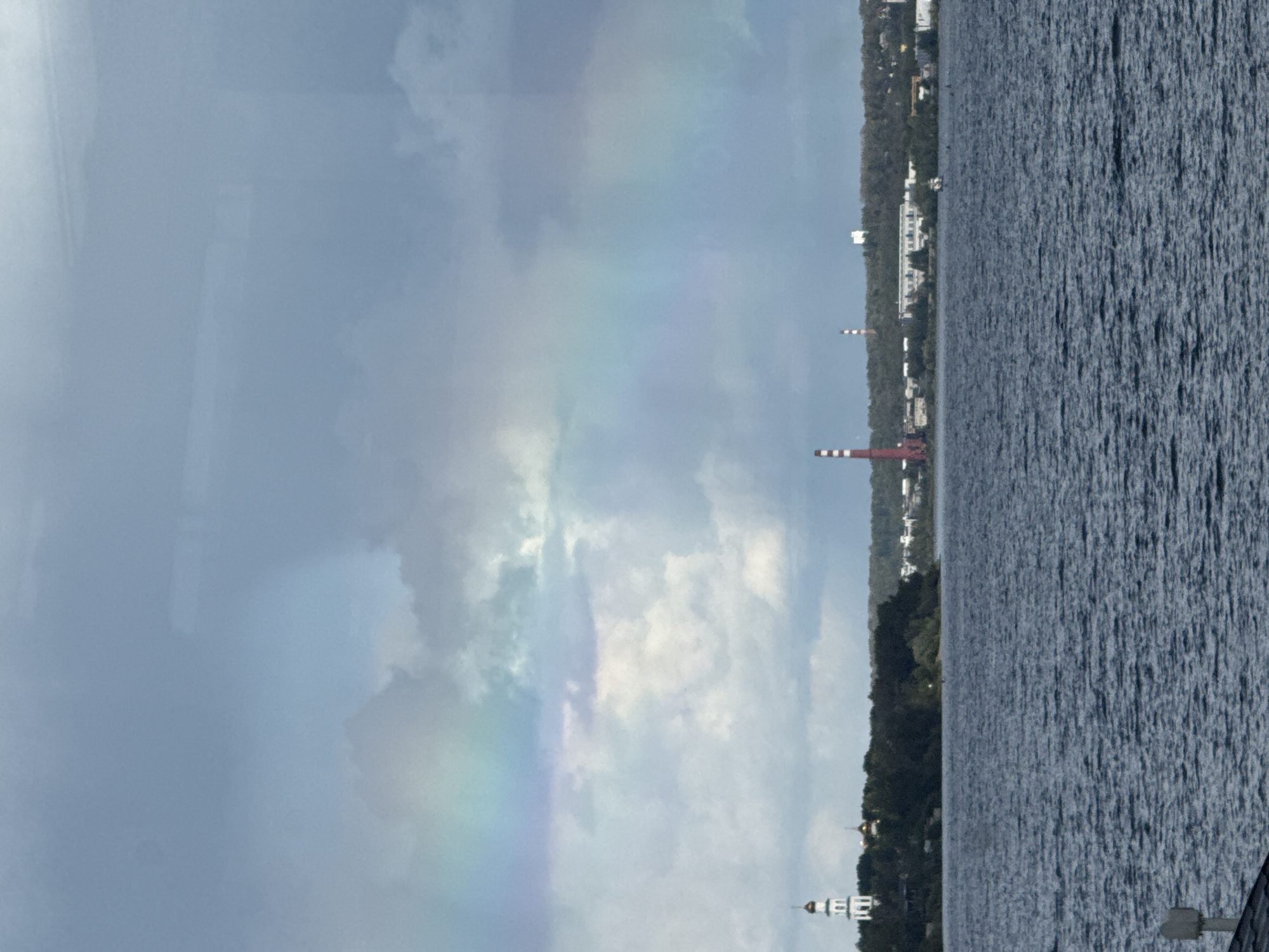 The image depicts a wide river in Perm, Russia, with a distant cityscape visible on the opposite bank. A faint rainbow arcs across a cloudy sky, suggesting recent or ongoing rain. On the left side of the shore, a white bell tower with golden domes stands out against a dense line of dark green trees. Further to the right, a tall red and white striped industrial chimney is prominent, accompanied by several low-rise buildings and what appears to be a power plant or factory complex. The water is a deep blue-grey with ripples, indicating a moderate current. A white structure, possibly a pier or part of a dock, is visible in the foreground on the lower left. The overall atmosphere is overcast and somewhat dramatic due to the rainbow and industrial elements.