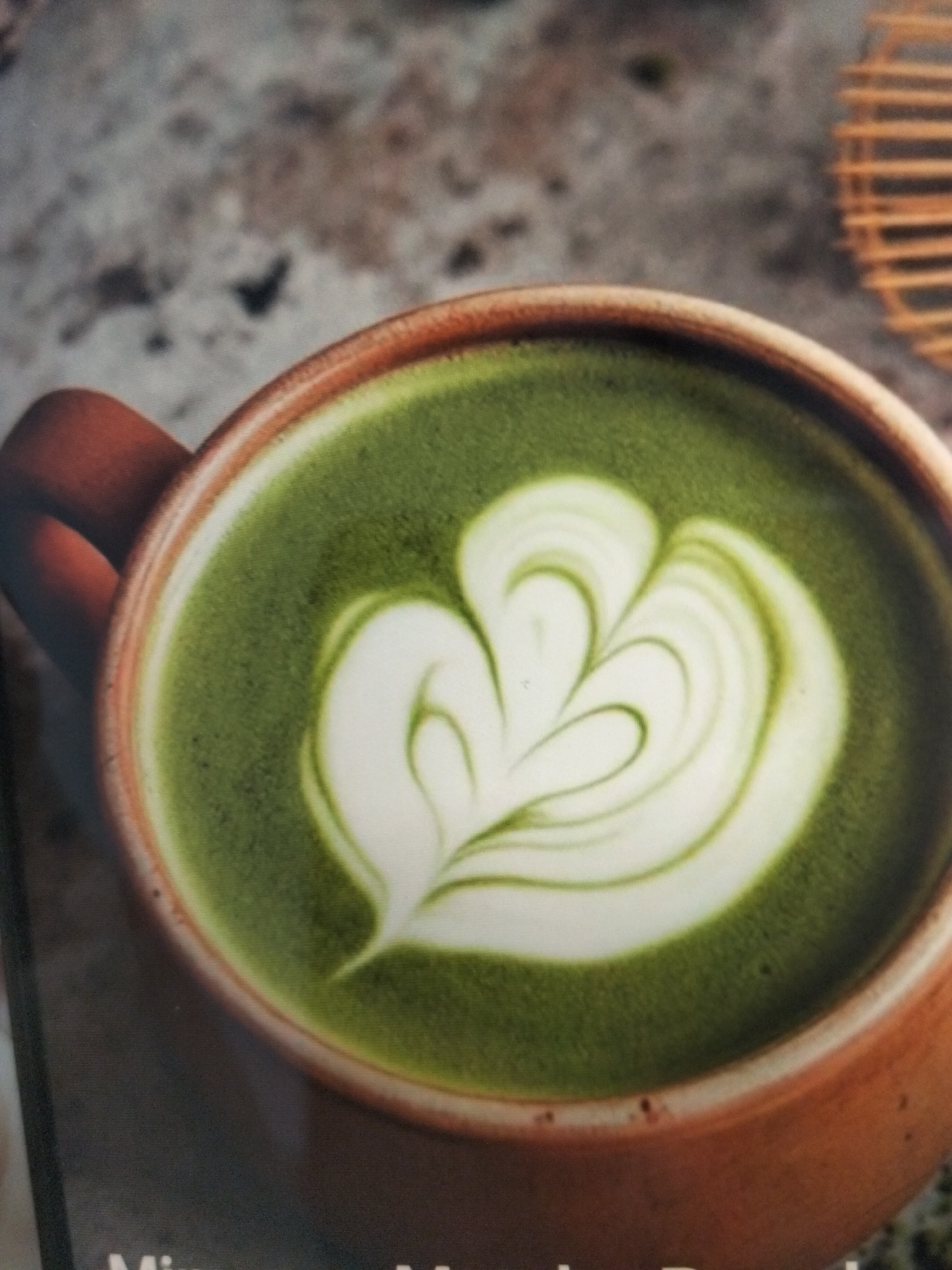 The image shows a close-up, top-down view of a mug of matcha latte. The latte has intricate white foam art resembling a stylized leaf or flower pattern. The mug itself is made of a reddish-brown ceramic material and has a handle visible on the left side. The background is a textured, speckled surface that appears to be a countertop or table, with a blurred wooden object with vertical slats visible in the upper right corner. There is text at the bottom of the image, but it is mostly out of focus. The lighting is soft, highlighting the details of the foam art. There are no people or any indication of the specific location in the city of Payagaboh, Indonesia, within the provided image.