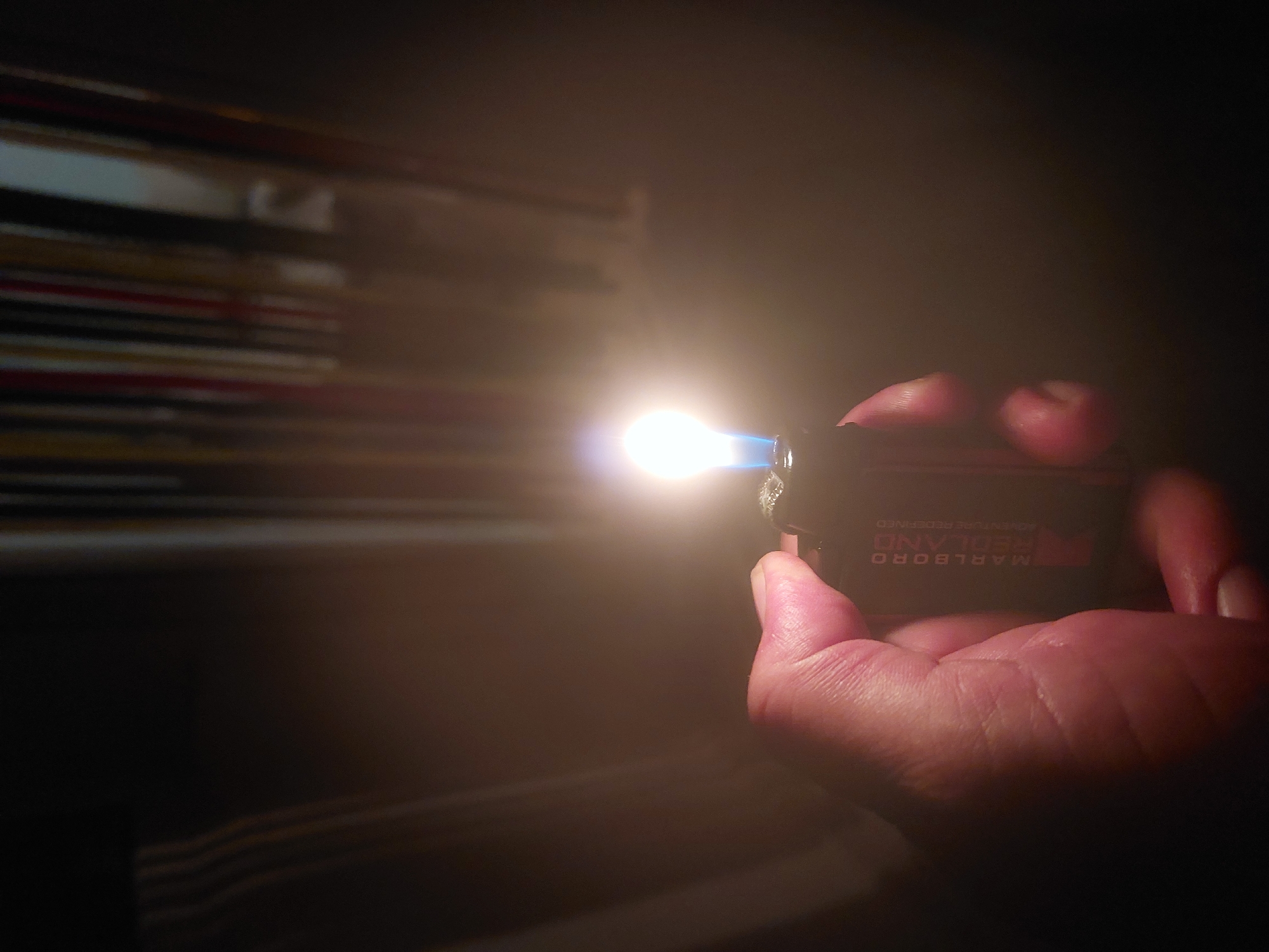 The image shows a hand holding a lit lighter in a dark room. The lighter is black and has the word "MARLBORO REDLAND" printed on it. The flame is bright and blue at the base, turning white towards the tip. In the background, there is a blurred object resembling a stack of papers or magazines. The lighting suggests the photo was taken in a dimly lit indoor setting. There is no information available to identify the location as Tsarevo, Bulgaria.