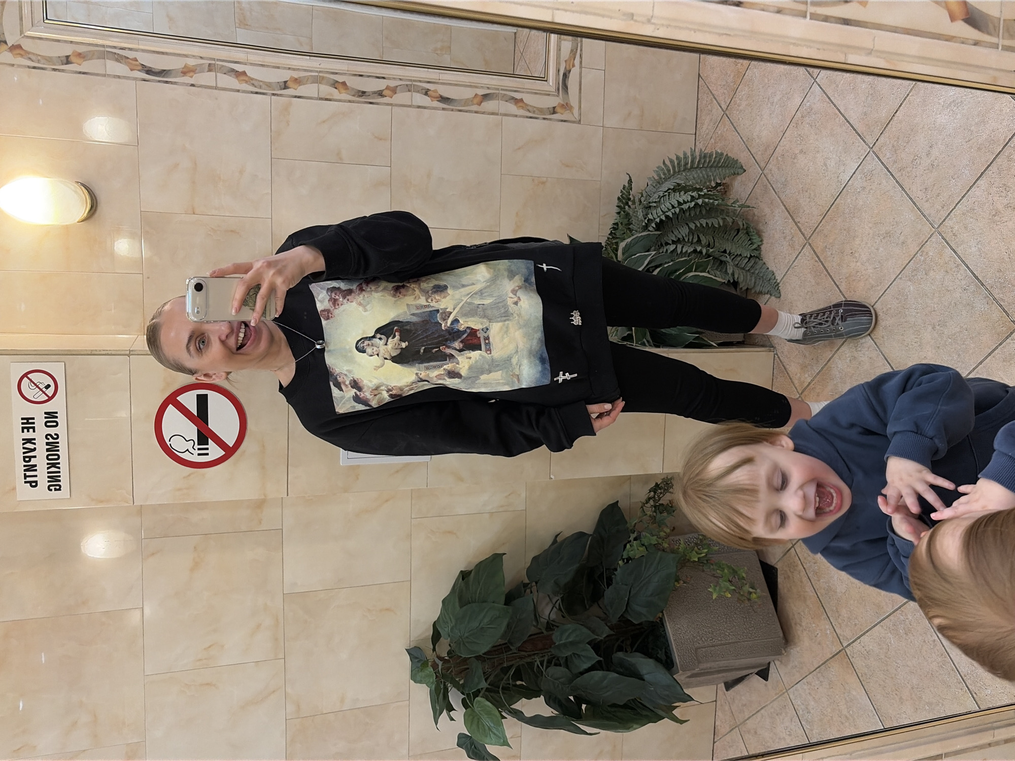 A woman and two small children are reflected in a mirror. The woman is holding a smartphone and appears to be taking a selfie. She is wearing a black sweatshirt with an image of a religious painting on the front, black leggings, and grey sneakers. One child is wearing a blue sweatshirt, laughing, and making a gesture with their hands. Only the head of the second child is visible. The background appears to be a bathroom with tiled walls and floor. There is a no smoking sign on the wall and potted plants on the floor. The style of the tiling around the mirror and light fixture suggest the bathroom may be in a public building. There are no elements in the image to indicate this location is specifically in Chaikovsky, Russia.
