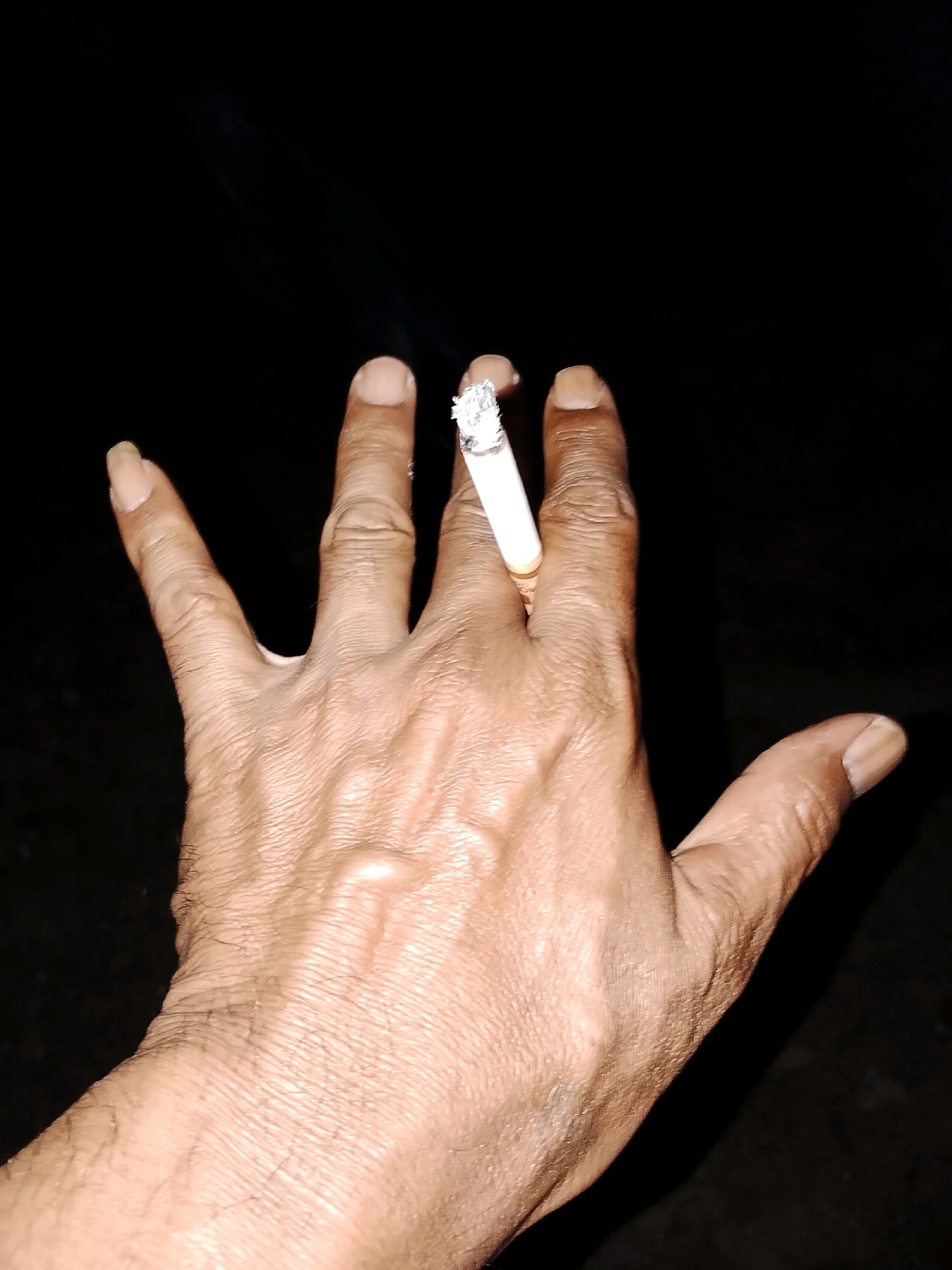 A close-up, eye-level, slightly tilted shot shows a hand holding a lit cigarette. The hand is positioned in the center of the frame, with the fingers splayed. The cigarette is held between the index and middle fingers, with the burning end facing upwards. Smoke is visible rising from the cigarette. The hand appears to be that of an adult male, with visible veins and skin texture. The background is dark and out of focus, suggesting an indoor setting or nighttime. There are no other people or objects visible in the frame.