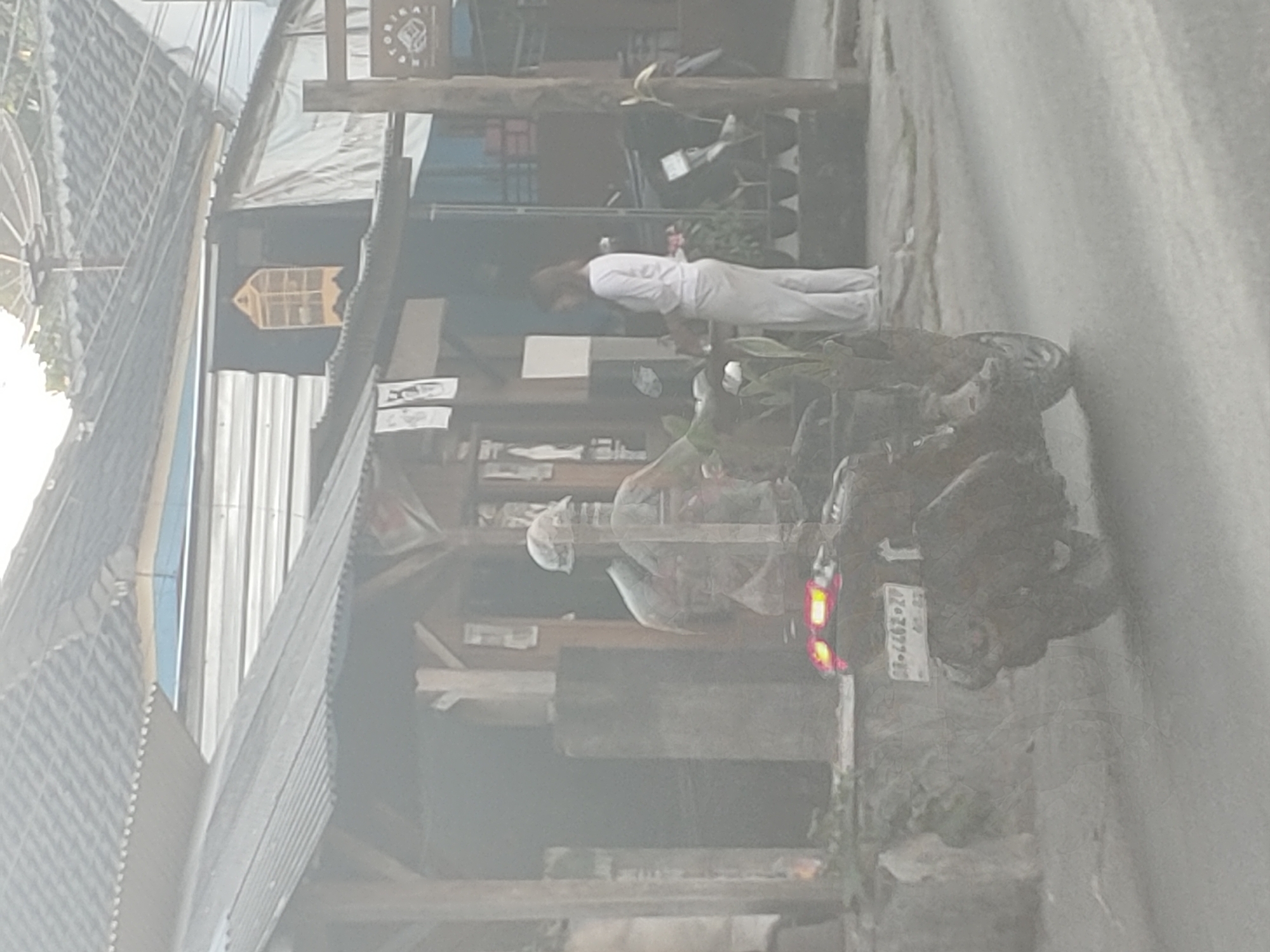 The image captures a street scene in Kembang Kerang, Indonesia. A person wearing a helmet is riding a scooter on a paved road. The scooter has a license plate that reads "BR 4462 ZP". Behind the rider, a woman in a white top and light-colored pants stands near a building. The building appears to be a shop or stall with wooden supports and a corrugated metal roof. Several potted plants are visible in front of the building. The overall atmosphere is that of a bustling street.