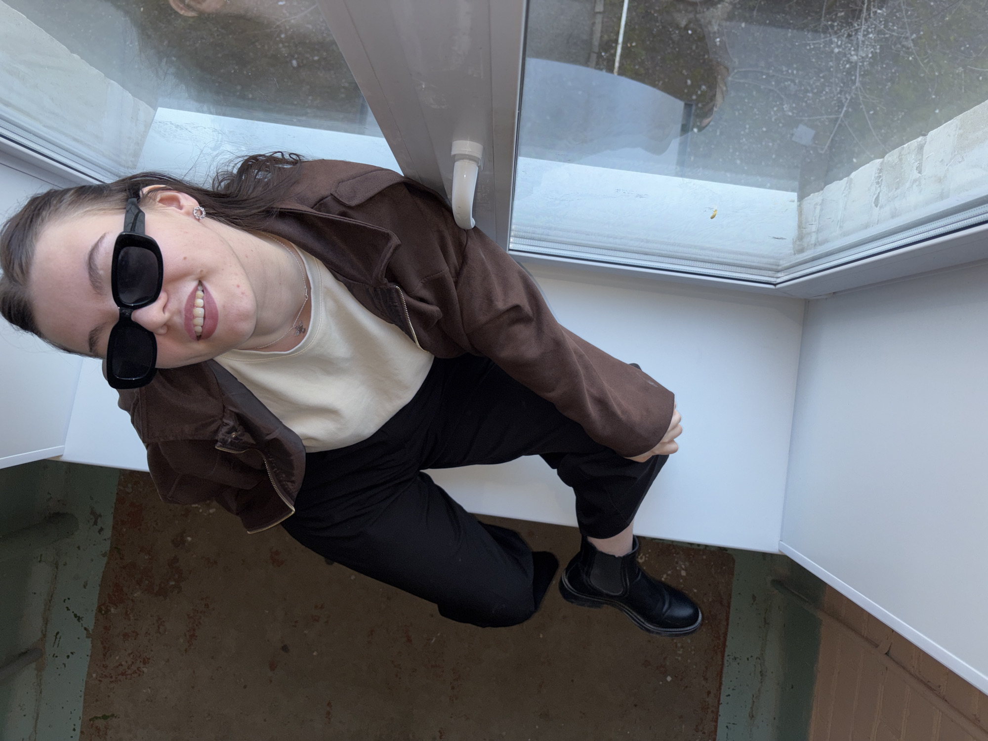 This image, taken from a high-angle perspective, shows a young woman looking up and smiling directly at the camera. She is positioned below a large, modern window with a white frame and handle.
The woman has dark hair and wears large, rectangular black sunglasses that cover her eyes. She is smiling genuinely, showing her teeth, and appears cheerful. Her attire includes a light cream or beige top, layered with a dark brown jacket or shacket, black trousers, and sturdy black Chelsea boots. Notable accessories include a silver flower-shaped stud earring in her visible left ear and a delicate silver necklace with a small pendant. Her left hand is resting on her knee, contributing to a relaxed pose.
The window through which the image is framed appears to be clean, with a white, possibly PVC, frame. The glass reflects a dull, overcast outdoor scene, showing what might be bare tree branches and a general lack of bright sunlight, suggesting daytime but possibly gloomy weather. There are some dust or water marks on the windowpane. Below the woman and the window is a ground surface that is rough and textured, with varying earthy tones of brown, green, and reddish-orange, indicating a possibly concrete or worn outdoor/industrial floor.
The scene conveys a casual portrait taken in daylight, with the woman as the central subject in a setting that combines modern architectural elements (the window) with a more utilitarian ground surface. The location context for this photograph is Perm, Russia. No text is visible within the image.