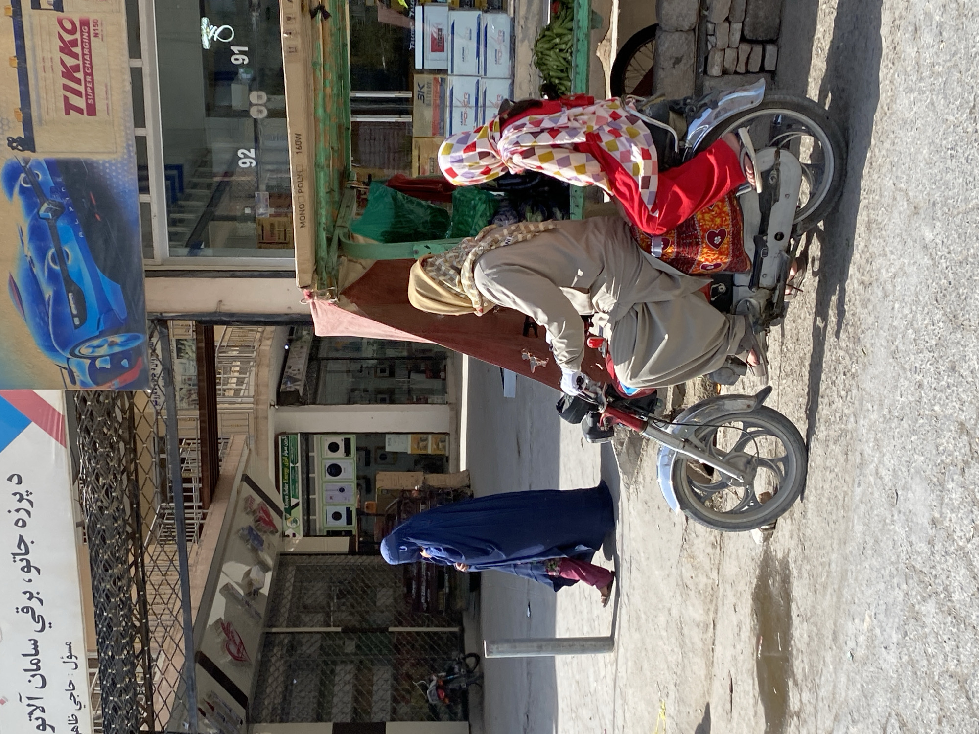 Two women are riding on a motorcycle in Kandahar, Afghanistan. The woman in front, wearing a beige headscarf and light brown clothing, is driving the motorcycle. The woman behind her wears a colorful headscarf with a checkered pattern and red pants. The motorcycle is parked on a dirt road in front of a market stall selling various goods, including batteries and what appear to be vegetables. In the background, a woman in a dark blue burqa walks away from the camera. The scene appears to be a typical street in a bustling Afghan city, with various shops and signs visible.