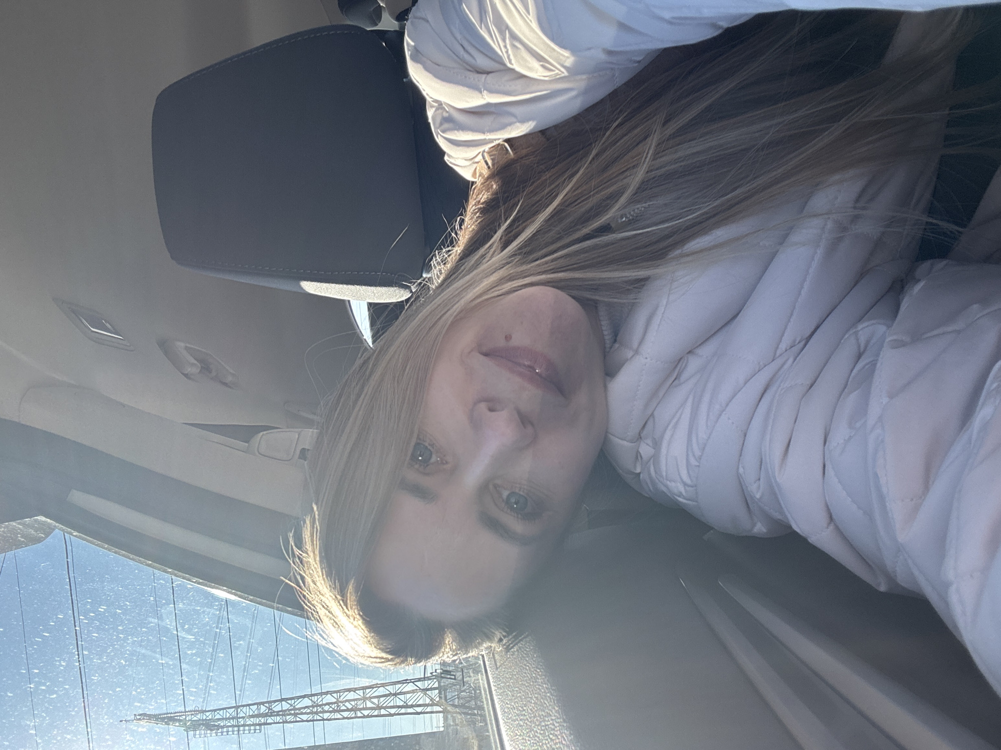A woman with blonde hair is shown in a selfie. She is wearing a white quilted jacket. The background shows the interior of a car with a grey headrest. Through the windshield, a bridge structure is visible, appearing to be the Kama Bridge in Chaikovsky, Russia.