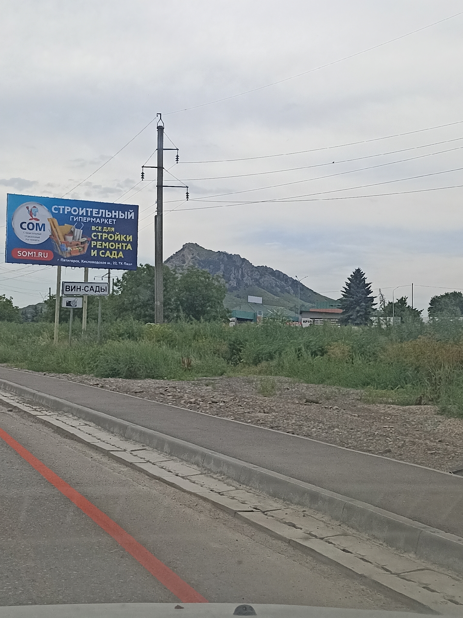 A wide shot shows a road in Vinsady, Russia, with a large billboard advertising a construction hypermarket. The billboard, featuring the "COM" logo and the slogan "Everything for construction, repair, and garden," is positioned to the left of the frame. To the right of the billboard, a sign reading "ВИН-САДЫ" (Vinsady) with a camera icon is visible. In the background, a mountain range dominates the landscape under a cloudy sky, with scattered trees and buildings. The road in the foreground has a red line painted on it, and a grey concrete curb runs alongside. The environment appears to be a semi-urban area with natural elements.