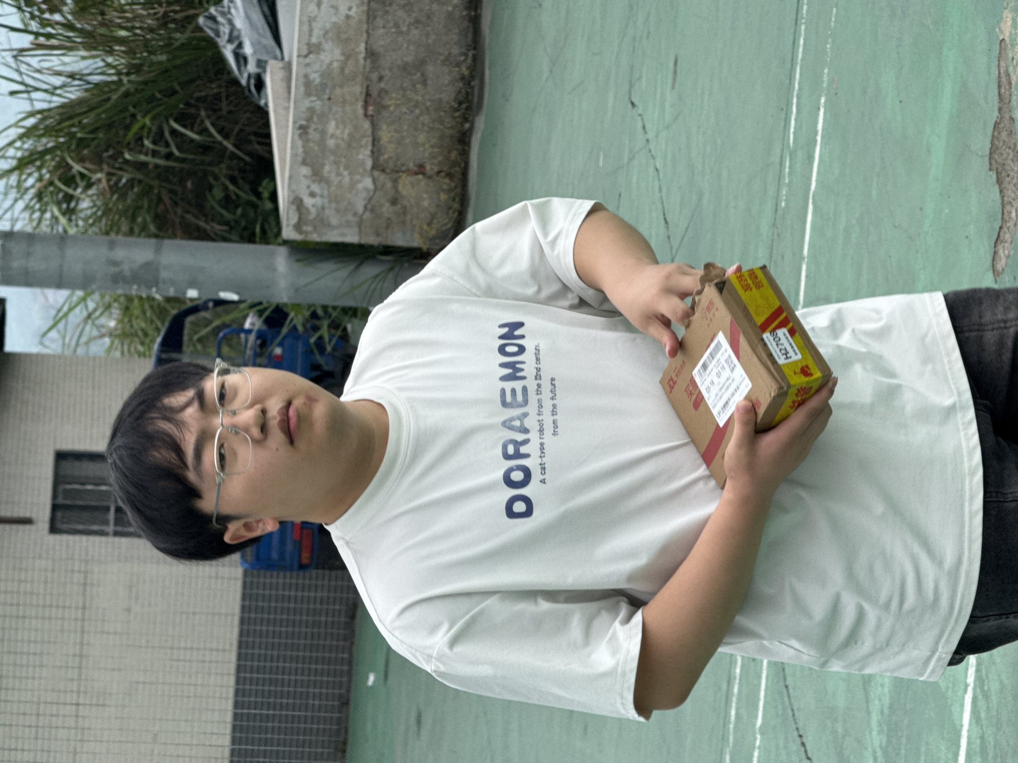 A person wearing glasses and a white t-shirt with the word "DORAEMON" on it is holding a cardboard box with Chinese writing on it. The writing appears to indicate a date of 03-18. The person is standing on a green surface with white lines, likely a court, outdoors in what appears to be a semi-urban or suburban area. A building is visible in the background with a barred window. There is also a utility pole, some vegetation, and a blue object that could be a vehicle. The location is Jiangjin, China.
