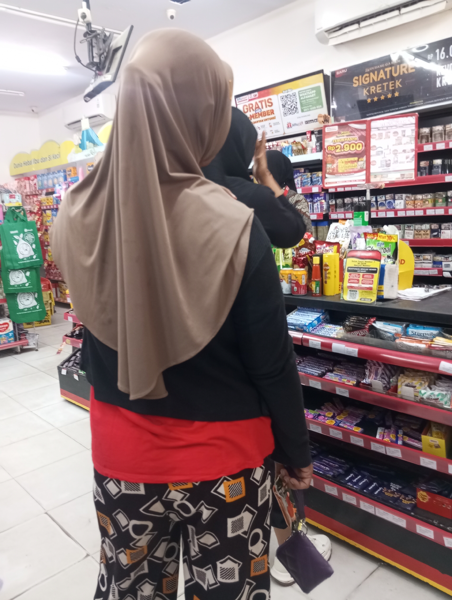 In the city of Tanjung Gading, Indonesia, two individuals are observed from behind in what appears to be a convenience store. One person, wearing a light brown hijab, a black long-sleeved shirt, and a red undershirt, stands in the foreground. They are also wearing patterned pants with a black base and white, yellow, and black geometric shapes. The second individual, behind the first, is partially visible and appears to be wearing a black top. They are carrying a purple pouch in their right hand.
The environment is a brightly lit retail space with shelves stocked with various products, including packaged food items, drinks, and cigarettes. Several signs are visible, including advertisements for "GRATIS MEMBER" (Free Member) and "SIGNATURE KRETEK" (Signature Clove Cigarettes). A security camera is mounted on the ceiling. The floor is tiled with light-colored squares. The overall scene suggests a typical transaction or browsing activity within a local store.