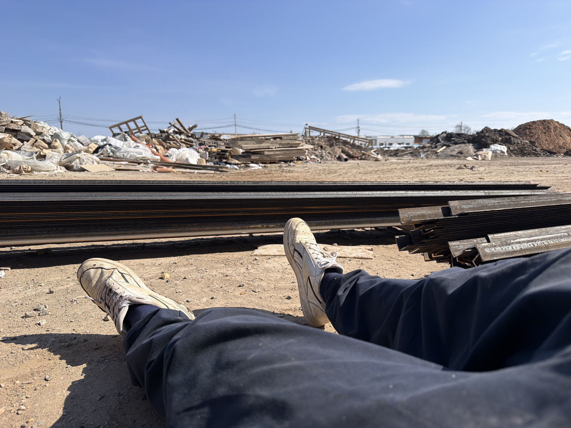 The image captures a first-person perspective of a person lying or sitting on dry, dusty ground at what appears to be an industrial or construction site in Chaikovsky, Russia. The legs of the individual are prominent in the foreground, extended forward. They are wearing dark blue trousers and white, somewhat soiled athletic shoes. A clear shadow is cast by the left leg and foot on the sun-baked earth, indicating bright sunlight.
In the mid-ground, a substantial stack of dark metal beams or profiles lies horizontally, forming a strong linear element across the frame. Beyond these materials, the background reveals a vast, open area covered with dry earth. Numerous piles of debris are scattered across the landscape; these include what looks like concrete rubble, discarded building materials, white sacks possibly containing waste, wooden frameworks, and mounds of reddish-brown soil or dirt.
In the distance, under a clear blue sky with a few faint clouds, faint outlines of buildings and power lines with poles are visible. The overall scene suggests an active or recently active industrial zone, possibly a demolition site, a storage yard for construction materials, or a landfill area. The time of day is bright and sunny, likely midday or early afternoon, given the clear shadows. No discernible text is visible in the image. The person's relaxed posture suggests a moment of rest or observation within this gritty, utilitarian environment.