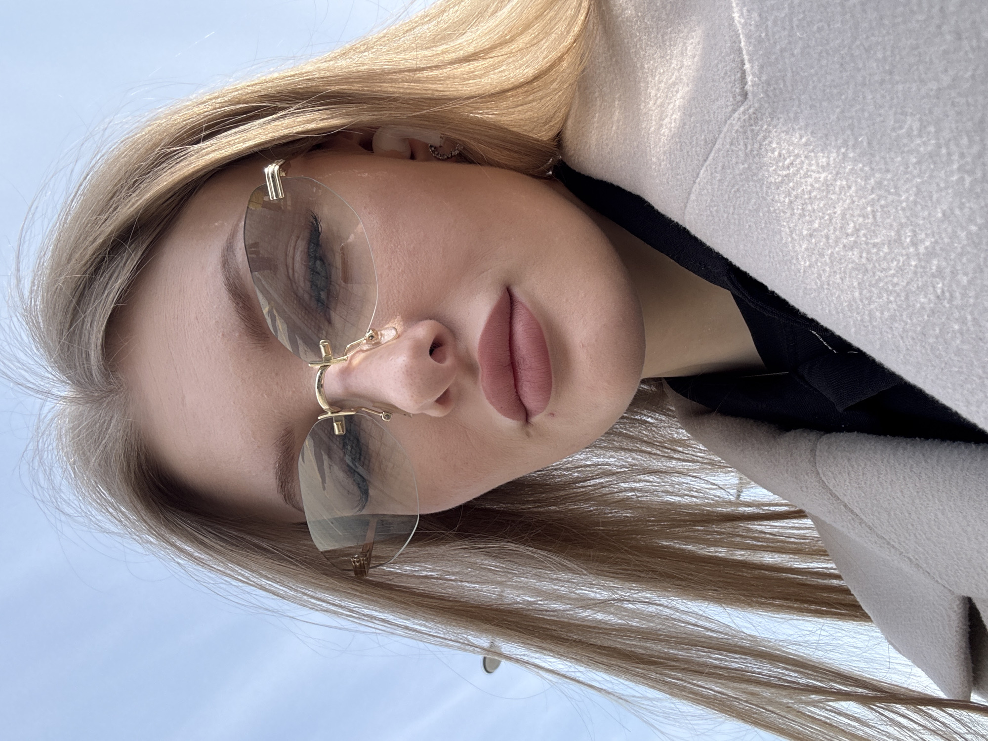 An outdoor, low-angle, close-up portrait depicts a light-skinned female individual with long, straight blonde hair falling past her shoulders. She wears rimless sunglasses with gold-colored temple and bridge accents, partially obscuring her eyes. Her lips are full and pink. A white wireless earbud is visible in her right ear, along with a small, heart-shaped earring. She is dressed in a light-colored (beige or off-white) textured coat over a dark-colored garment. The background consists of a clear, light blue sky, with a dark streetlamp partially visible in the upper left. The scene is located in Perm, Russia.