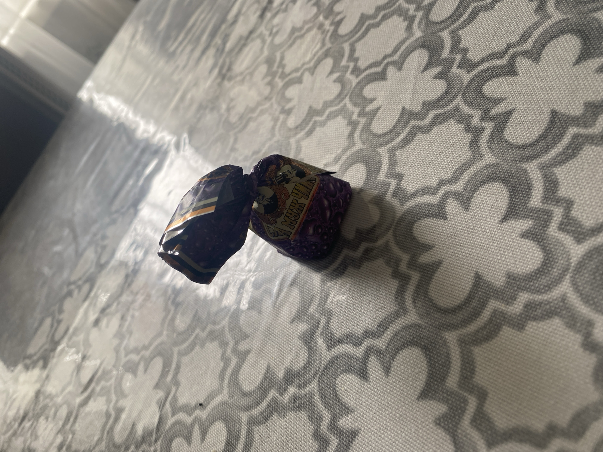 A candy wrapper is seen lying on a patterned surface. The wrapper is predominantly purple with some black, orange, and white accents. There is text and images visible on the wrapper, including what appears to be a man in a top hat. The patterned surface is light-colored with a repeating grey design. The scene is lit by a bright light source casting shadows. The image appears to be taken indoors, on a table or similar flat surface. There are no people or specific city landmarks visible in the image.