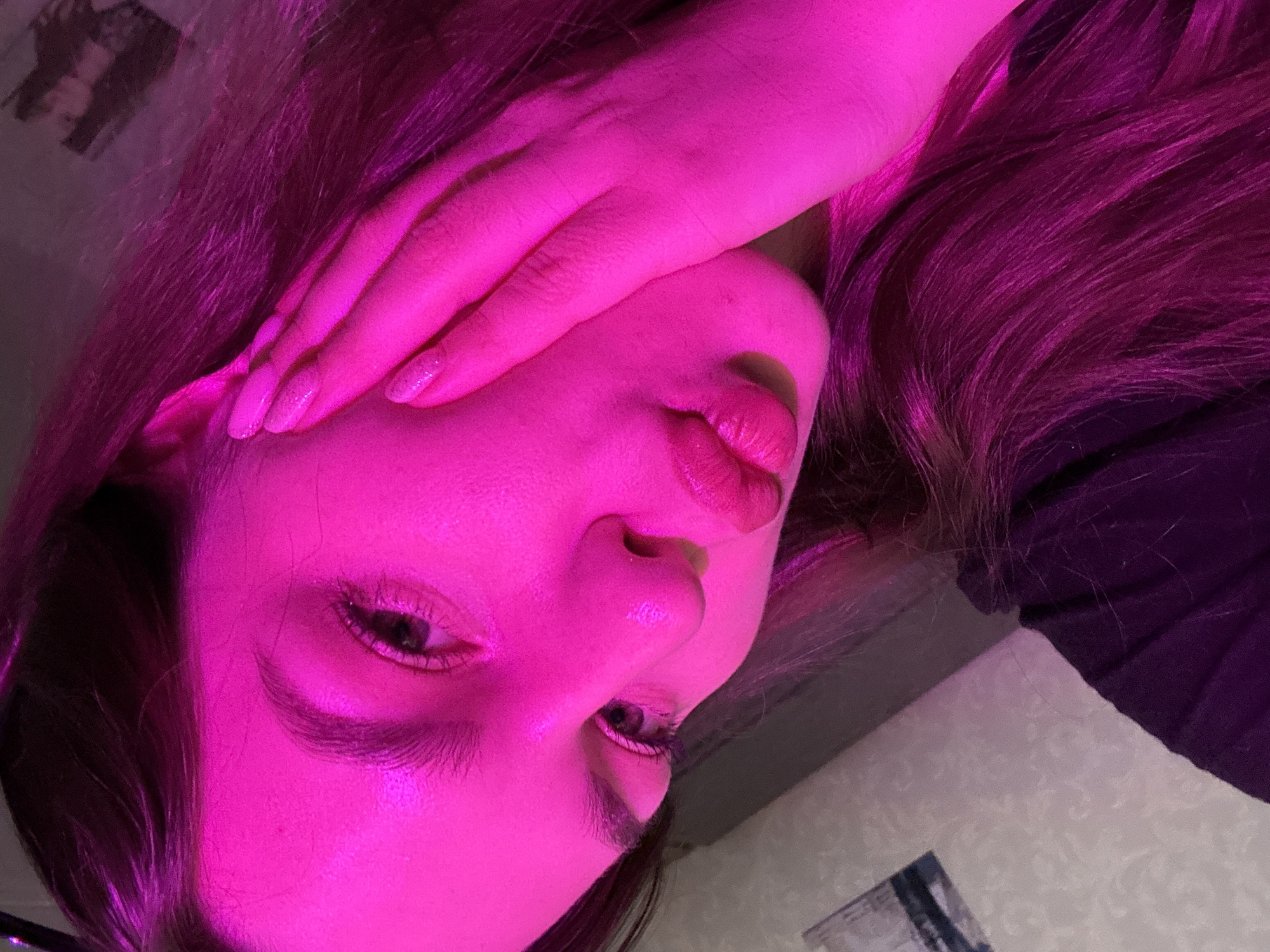 The image is a close-up, waist-up shot of a young woman illuminated by pink lighting. She is looking down and to her left, with her right hand touching her cheek. Her lips are pursed. The background appears to be a room with patterned wallpaper and what looks like a framed picture on the wall. There is no indication of the city of Chaikovsky, Russia, or any specific location details in the image itself. The woman has dark hair and is wearing a dark garment. The overall impression is of a selfie or portrait taken in a dimly lit environment with colored lighting.