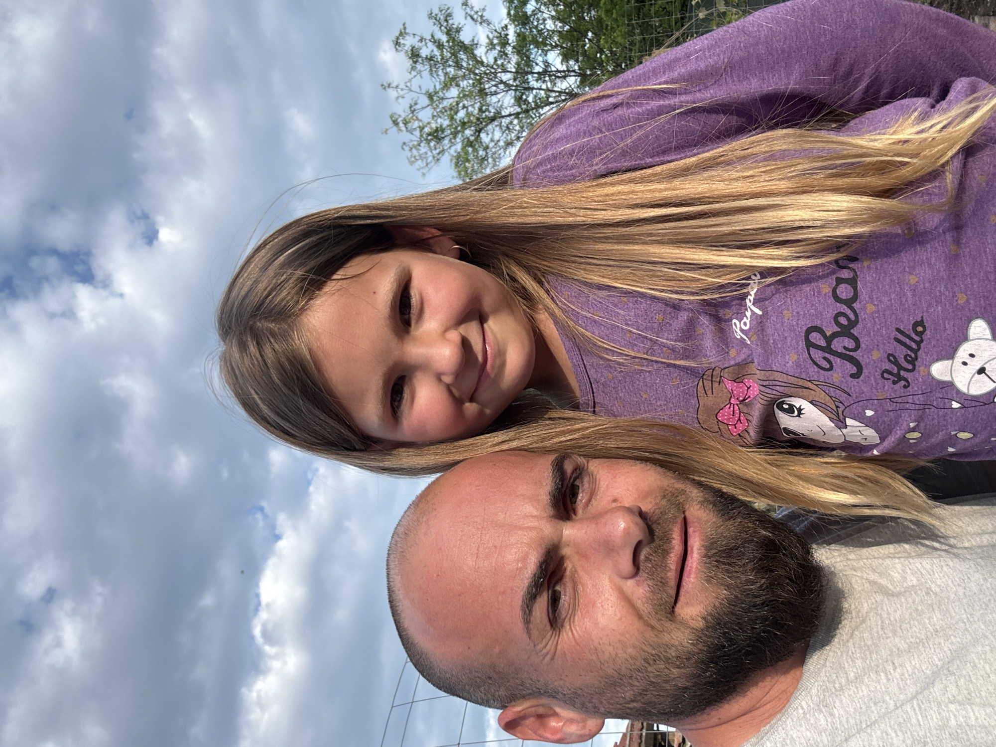 A selfie-style image shows a man and a young girl outside. The man has a shaved head, a dark beard, and is wearing a gray t-shirt. The girl has long blonde hair and is wearing a purple long-sleeved shirt with a cartoon print on it. They are positioned close together, with the girl above and slightly behind the man. The background features a cloudy sky, tree branches, and what appears to be a wire fence. The location is Sarnevo, Bulgaria, as indicated by metadata.