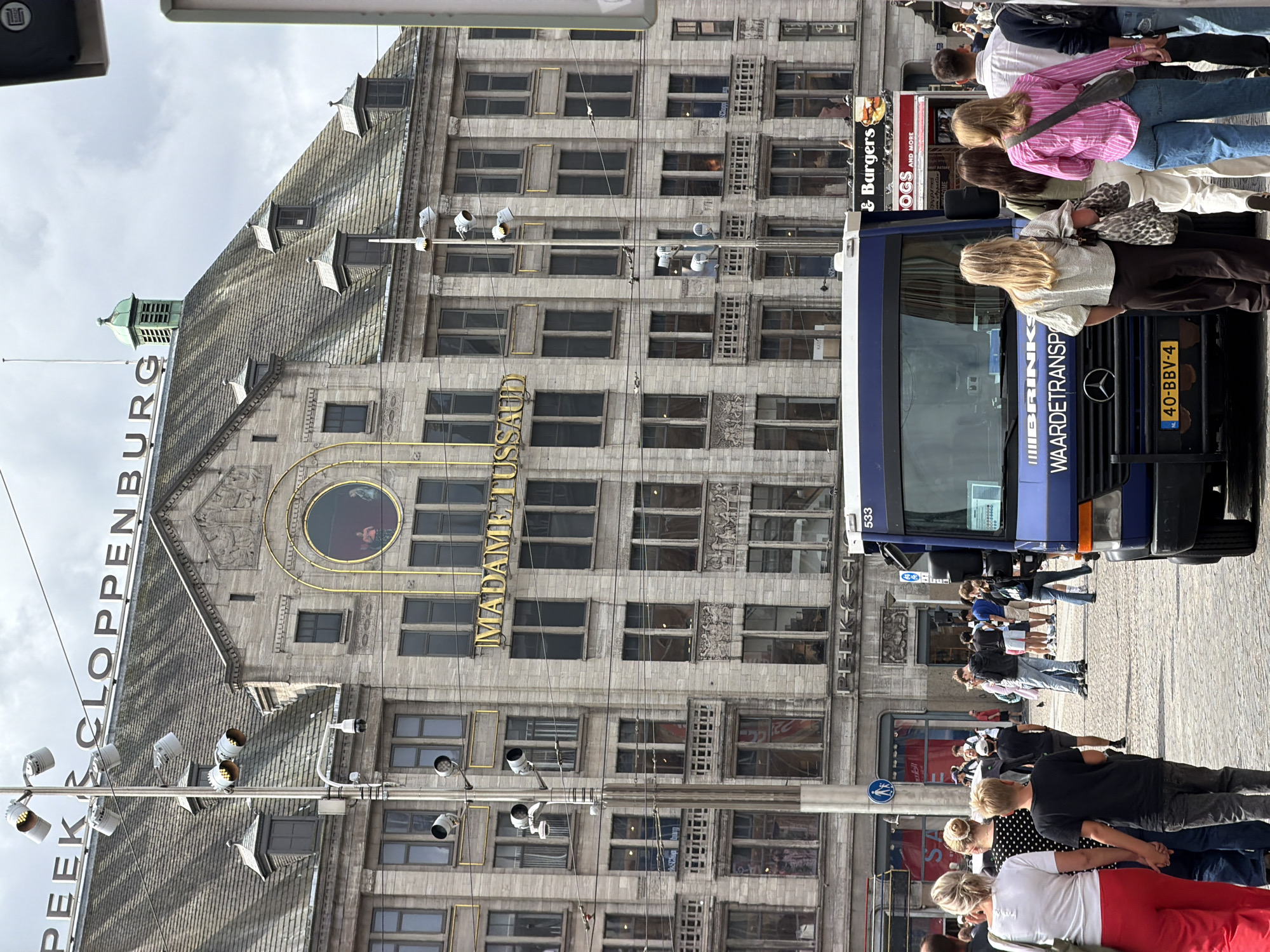 The image captures a street scene in Amsterdam, Netherlands, featuring the iconic Madame Tussauds wax museum. The building facade, constructed of light-colored stone, displays classical architectural elements with multiple rows of windows. A prominent sign above the entrance reads "MADAME TUSS AUD." On the rooftop, a green, louvered cupola is visible, along with multiple streetlights and a security camera mounted on a pole. In the foreground, a blue utility van with the text "BRINKS WAARDETRANSPORT" is parked, its license plate reading "40-BBV-4." Several people are walking on the cobblestone street, some in small groups, and others individually. A woman in a white top and red pants is walking away from the camera, holding hands with a young boy in a black t-shirt. Another woman in a polka-dot top and jeans is beside them. The background shows more of the building's facade and other establishments, including a sign for "Burgers." The sky is overcast with scattered clouds.
