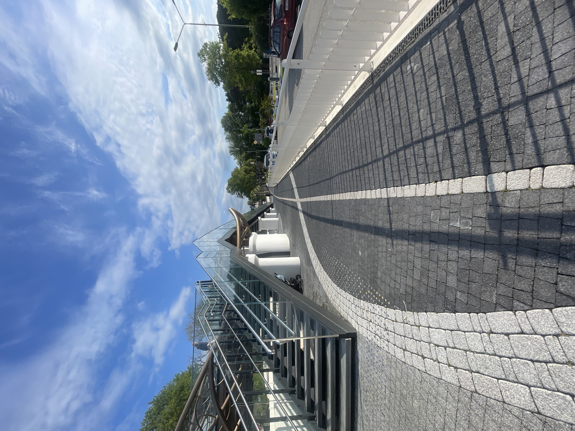 The image depicts a pedestrian walkway in Sochi, Russia. The walkway is paved with dark gray cobblestones, bordered by white stones on the right side, creating a curved design. A modern staircase with glass walls and a metal railing leads to an elevated platform or structure. The sky is partly cloudy with a mix of blue sky and white clouds. Trees and vehicles are visible in the background to the right, suggesting a road or parking area. A white fence runs parallel to the walkway. The scene appears to be taken in daylight, likely during the afternoon. A shadow from the fence falls across the cobblestone walkway.
