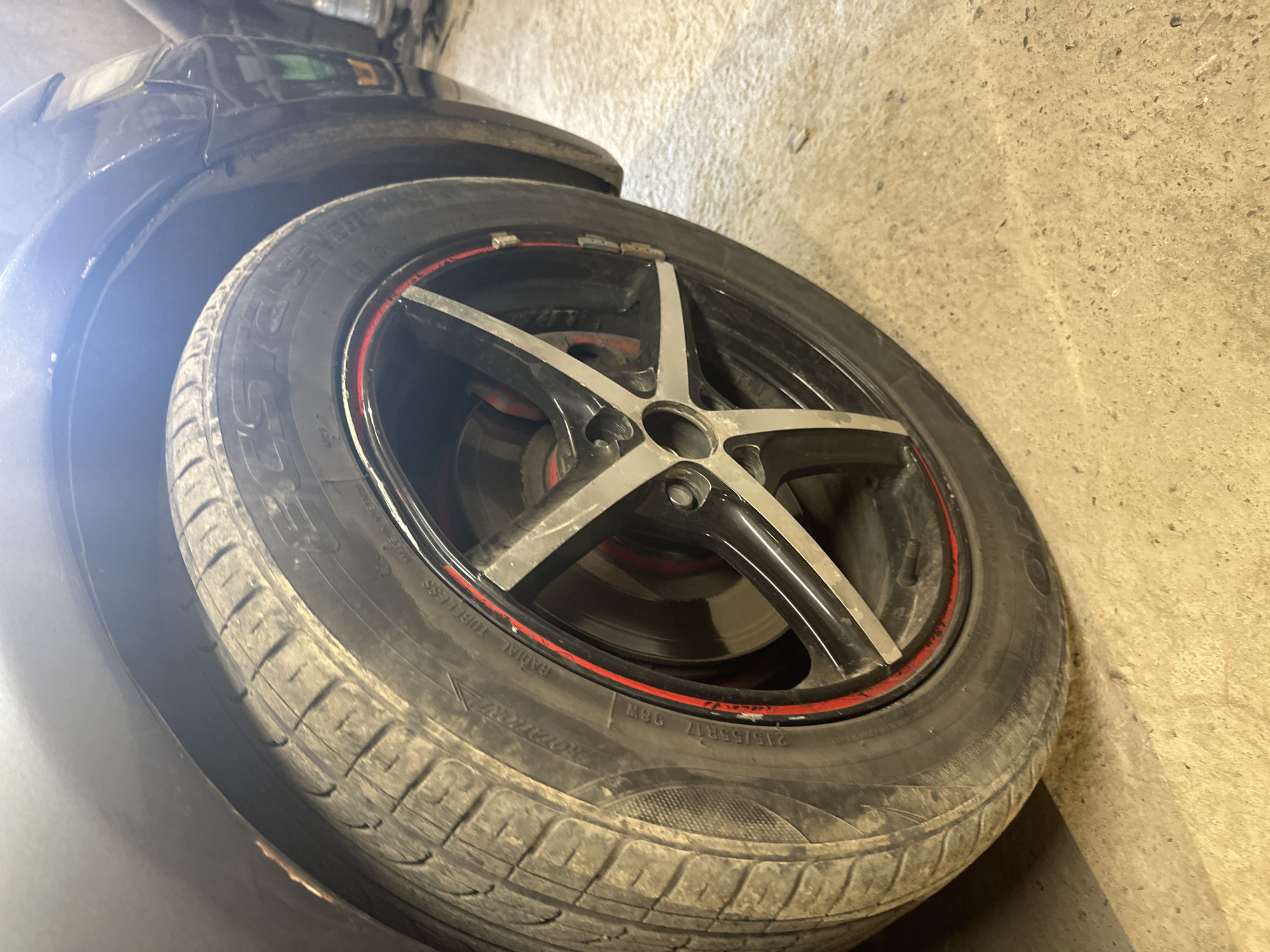 In the city of International, Kyrgyzstan, a close-up, eye-level shot captures the front passenger-side wheel of a dark-colored car, possibly a sedan, resting on a concrete floor. The tire is a Rotational Radial tubeless tire with the size marking "215/55R17 98W" visible. The wheel is a black alloy rim with silver accents and a red stripe around its circumference. The tire itself appears dusty and moderately worn. Part of the car's fender and bodywork is visible in the upper left and right portions of the frame, with the background showing a dimly lit, possibly industrial or garage-like environment. The lighting suggests the image was taken indoors, with artificial light casting reflections on the car's paint and the concrete floor. No people or other vehicles are present in the frame.