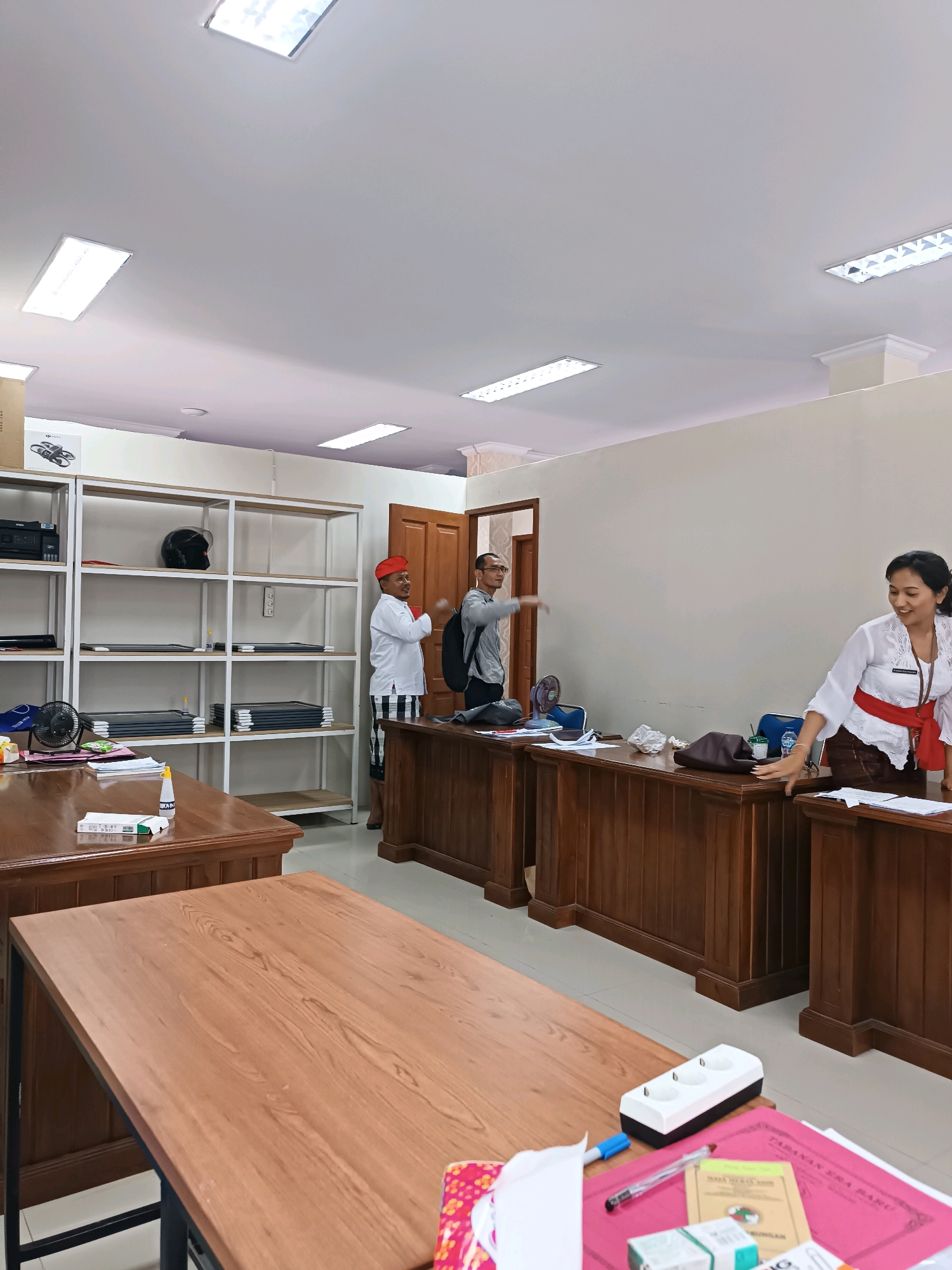 The image depicts an indoor scene, likely an office or administrative building in Tabanan, Indonesia. The room features several wooden desks arranged in a way that suggests a reception or service area. Behind the desks, a large shelving unit is visible, stocked with various items including boxes, what appears to be a helmet, and stacks of documents or folders.
Three individuals are present. On the left, a person wearing a red head covering and a white shirt with patterned pants stands near the shelving unit. In the center, a person in a grey shirt and dark pants, wearing glasses, is standing behind a desk, appearing to be in motion. On the right, a woman in a white top with a red sash and brown skirt stands behind a desk, smiling and looking towards the center of the room. She is reaching out with her right hand.
The environment includes fluorescent ceiling lights, a light-colored wall, and tiled flooring. Various office supplies are visible on the desks, including papers, pens, a power strip with multiple outlets, and a bottle of liquid. The overall impression is one of a functional administrative space.