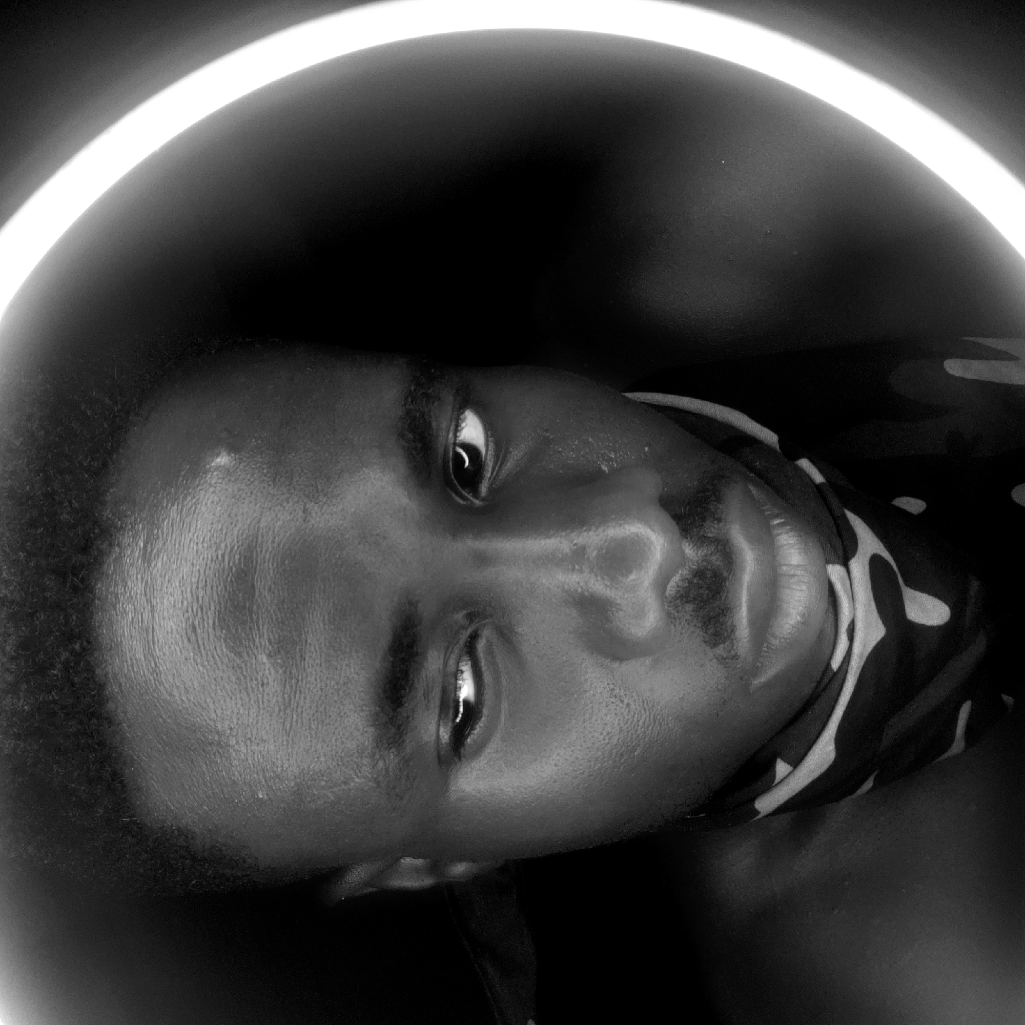 A close-up, black and white portrait of a young Black man is centered within a luminous ring light. The man's face is slightly tilted, with his eyes partially open and directed toward the viewer. His skin is smooth, and his hair is closely cropped. He appears to be wearing a patterned garment around his neck. The background is dark and out of focus. There is no indication of the location being Narayi, Nigeria.