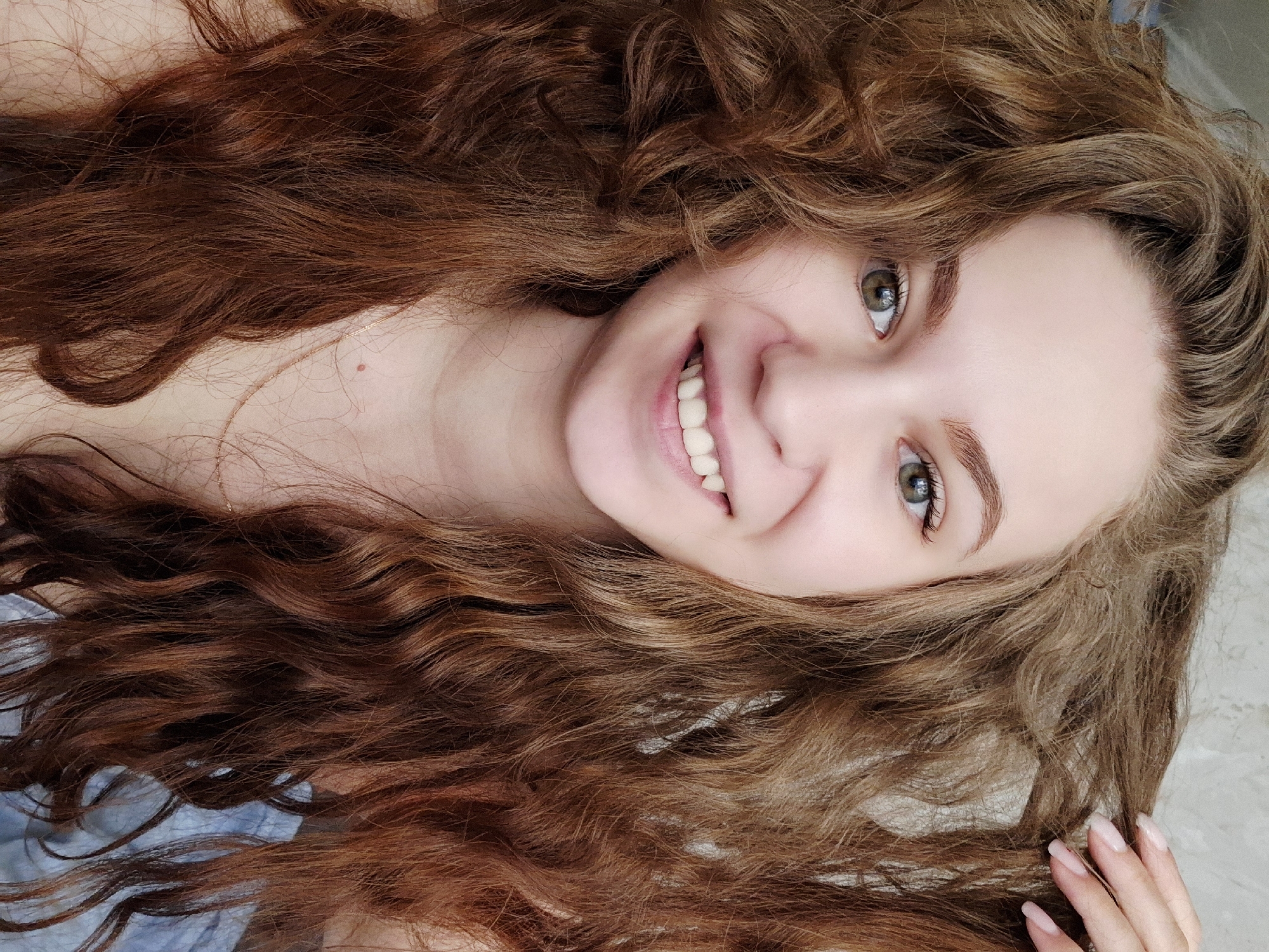 A young woman with long, curly, reddish-brown hair lies on a gray, textured surface. She is smiling, exposing her teeth. Her eyes are light green. A thin necklace is visible on her neck. Her right hand rests on the gray surface, with long, manicured nails. A blue fabric is visible beneath her left shoulder. The location is Chaikovsky, Russia.