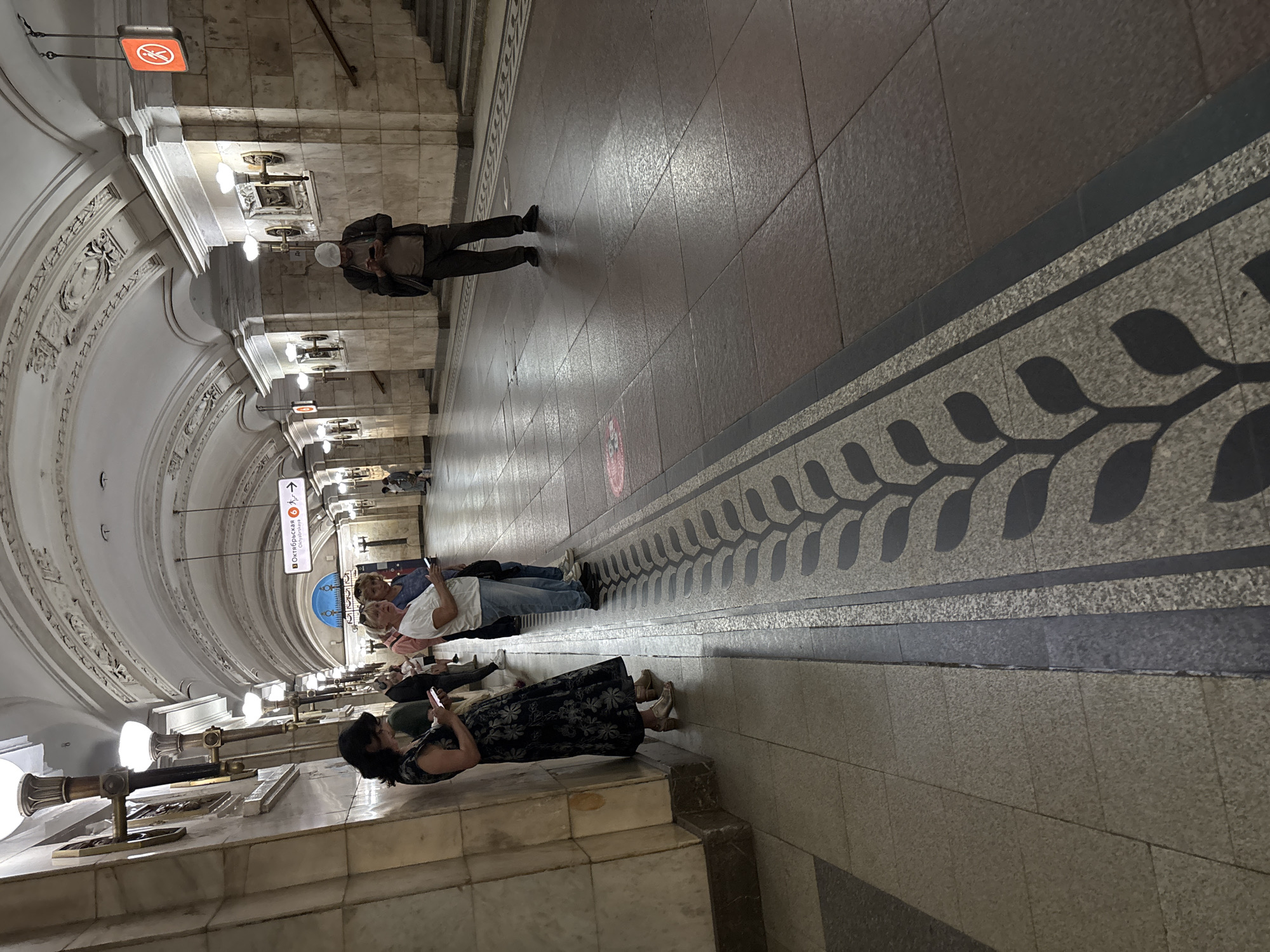 The image shows a well-lit, ornate metro station concourse in Moscow, Russia. The ceiling is a vaulted arch decorated with stucco reliefs. The walls are clad in polished stone with decorative sconce-style light fixtures. A decorative patterned border runs along the floor at the base of the wall, featuring a repeating leaf motif.
In the foreground left, a woman in a dark floral dress and gold heels is leaning against the wall, looking down at her phone. To her right, three people stand facing away from the camera, also looking at their phones. Further down the platform, a man in dark trousers, a light brown t-shirt, and a dark jacket, wearing a white cap, stands in the middle of the walkway, engrossed in his phone. The floor is tiled with large granite slabs. Signs are visible in the background, including a red circular sign with a pedestrian symbol. The station appears clean and well-maintained.