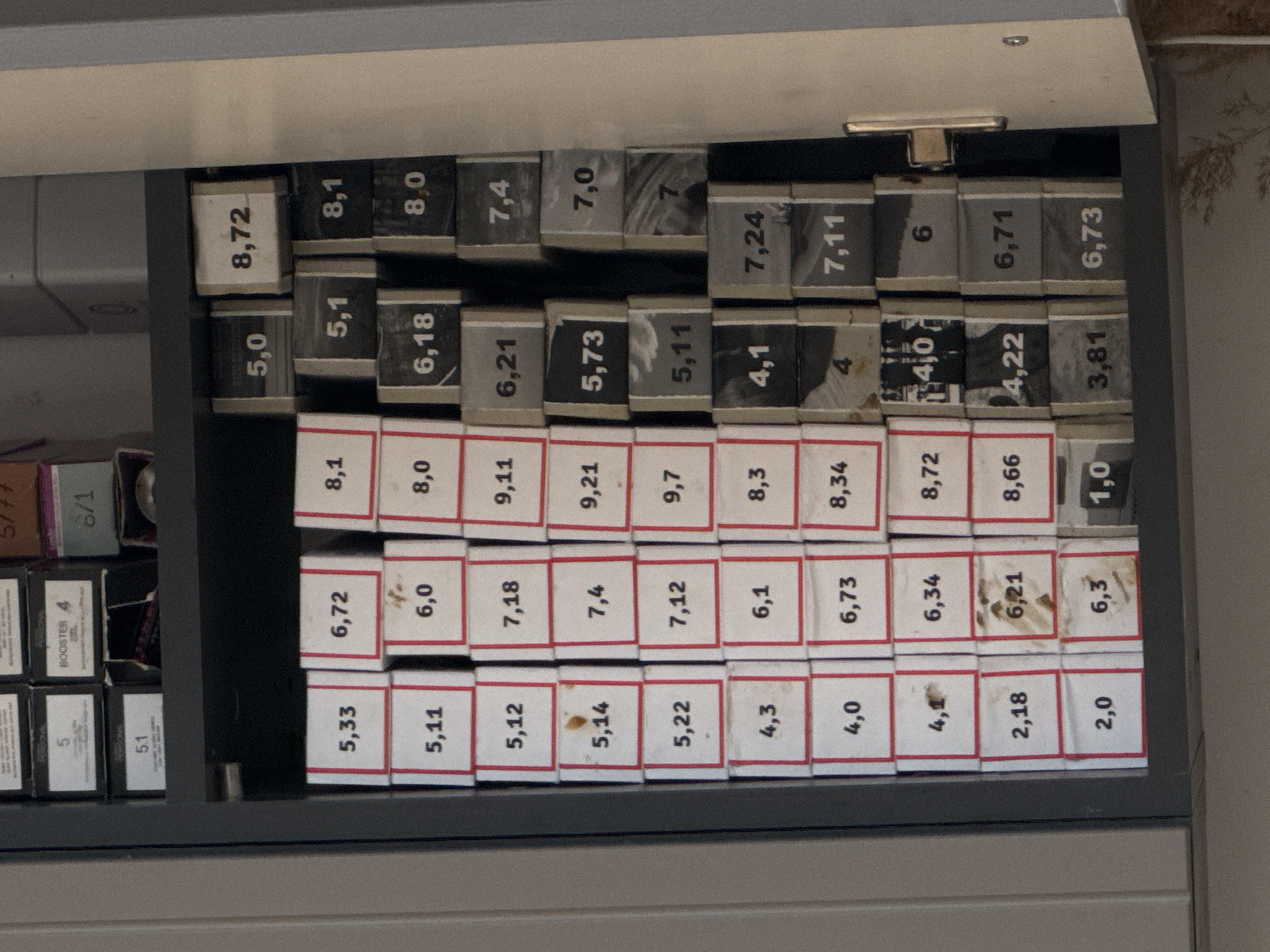 The image shows a close-up of a cabinet drawer filled with small boxes. The boxes appear to contain hair dye or similar products, judging by the numerical codes printed on them such as "8,72", "5,0", "5,33", "6,72", "8,1", "5,1", and many others. The codes vary, suggesting different shades or formulas. The boxes are arranged in neat rows and columns within the drawer. The drawer itself is part of a gray or dark-colored cabinet. There is some wear and tear visible on the boxes.