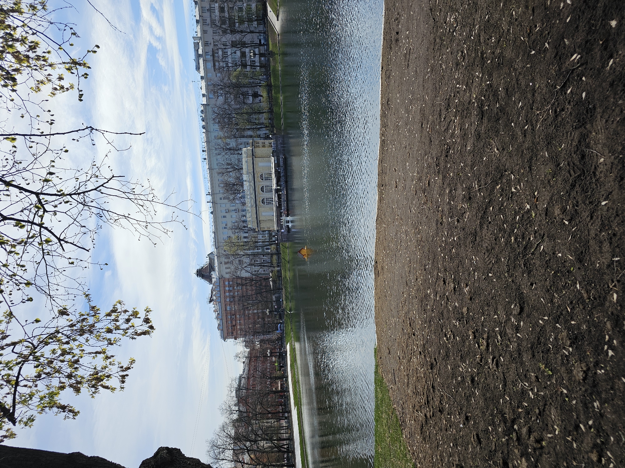 The image shows a cityscape with a pond in Moscow, Russia. The scene is taken from ground level, with a dirt path on the right and tree branches with budding leaves on the left. In the middle, a body of water, likely a pond or river, reflects the sky and buildings on the opposite bank. The architecture ranges from older, ornate buildings to modern apartment blocks, suggesting a blend of historical and contemporary elements. A small, possibly yellow, object floats on the water's surface. The sky is partly cloudy, indicating a pleasant day.