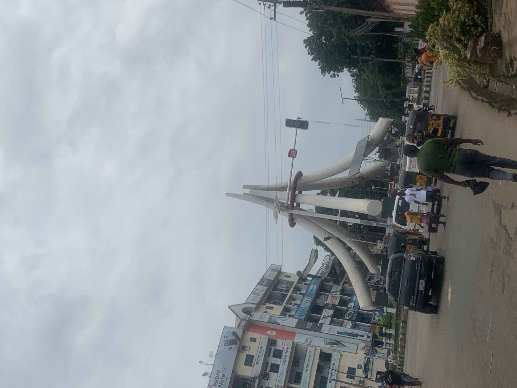 The image captures a street scene in Onitsha, Nigeria, featuring a prominent roundabout with a distinctive monument. The monument consists of several large, curved white structures that converge at the top, resembling abstract arms reaching upwards. In the background, a multi-story building is visible, adorned with numerous advertisements, primarily for "REDMI Note 15" and "TECNO." A traffic light with a digital timer displaying "58" is positioned above the roundabout. Several vehicles are present on the road, including a black pickup truck, a yellow and black auto-rickshaw, and a white vehicle. People are also visible, some walking and others in or around the vehicles. The environment appears to be a busy urban intersection under an overcast sky.