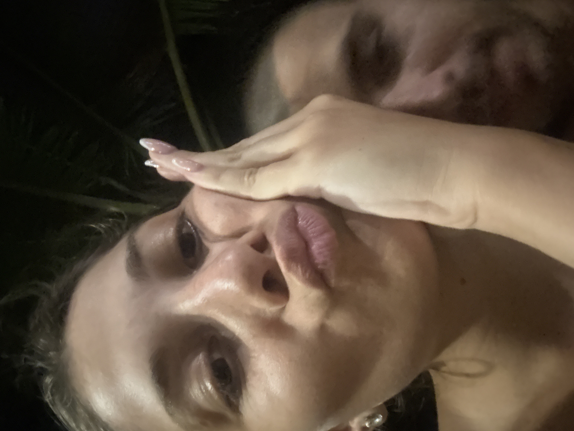 A selfie-style photograph depicts a woman with fair skin, light hair, and manicured nails posing with a pouting expression. Her hand covers one eye, while the other is visible. A second person is partially visible in the background, out of focus. The background is dark and appears to be outdoors, with some foliage visible. It is impossible to determine the location in Đà Nẵng, Vietnam, from the image.