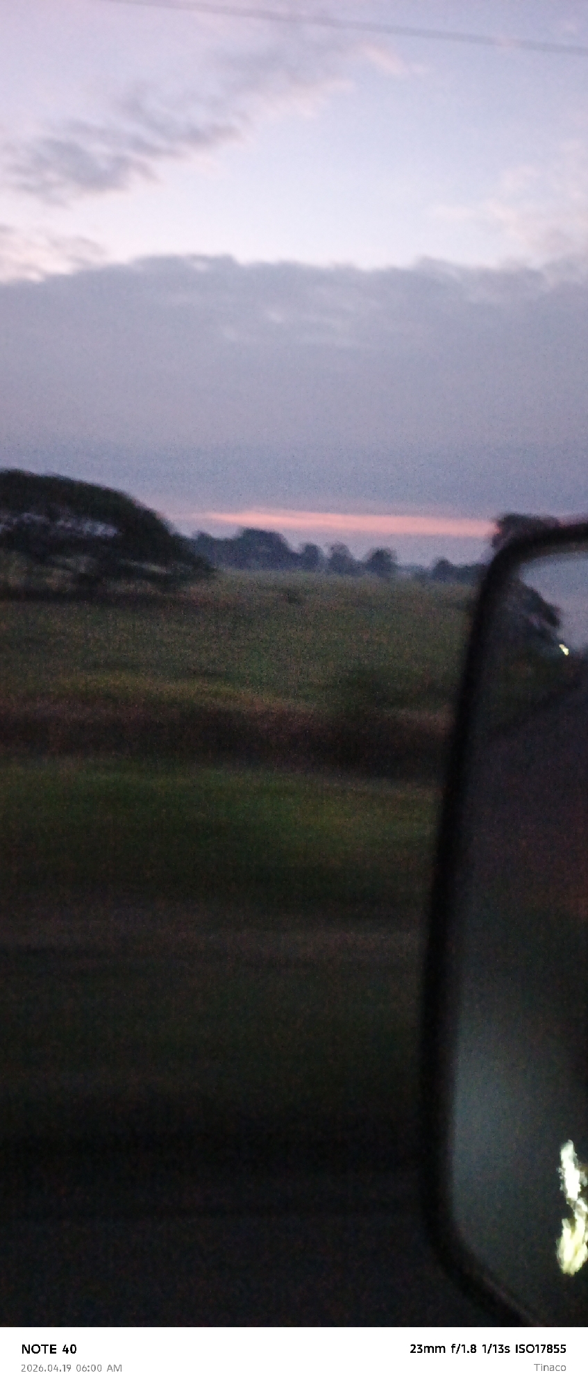 The photograph, taken in Tinaco, possibly near Cano Negro, Venezuela, at 6:00 AM on April 19, 2026, displays a landscape at dawn or dusk, viewed from inside a vehicle, evidenced by the side mirror visible on the right. The sky exhibits a gradient of pastel colors, transitioning from a light pinkish-orange near the horizon to deeper shades of purple and gray above, suggesting either sunrise or sunset. Below the horizon is a blurred view of a field or grassy area, possibly agricultural, with dark green patches interspersed with lighter areas. A line of trees or shrubs is silhouetted against the horizon, adding depth to the scene. The image exhibits motion blur, indicating that the photograph was taken while the vehicle was in motion. The camera settings, as indicated in the lower right corner, include a 23mm lens, an aperture of f/1.8, a shutter speed of 1/13s, and an ISO of 17855. The composition is unbalanced, with the foreground dominated by the dark interior of the vehicle and the side mirror. The overall ambiance is dim and muted due to the low light conditions.