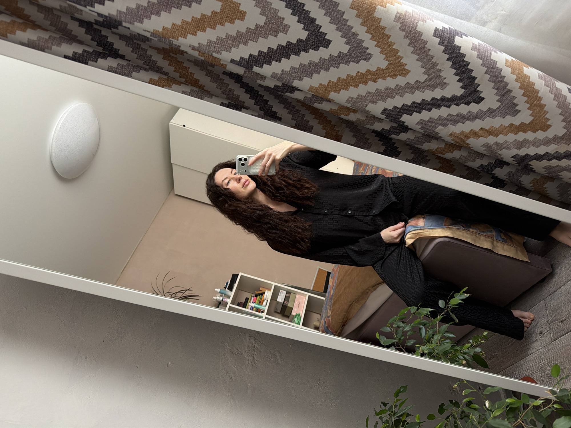 This image depicts a full-length mirror selfie taken by a female subject within a domestic interior in Perm, Russia. The subject, a woman with long, dark, wavy hair, is observed in the reflection. She wears a black, textured or subtly patterned two-piece pajama set, consisting of a long-sleeved button-down shirt and trousers. Her bare feet are visible. She is seated on what appears to be a dark brown sofa or bed frame, with her right leg extended and her left leg bent. She holds a light-colored smartphone in her right hand, positioning it to capture her reflection. Her gaze is directed towards the phone or mirror.
The reflected environment reveals several details:
To the left of the subject, a white shelving unit is visible, holding various items including bottles, boxes, and what appear to be books or files. Below the shelves, a section of a bed with patterned bedding in shades of brown, blue, and white is seen.
Above the subject, a round, white, flush-mounted ceiling light fixture is present.
To the right of the subject, partially reflected, and also visible directly outside the mirror frame on the right side of the image, are curtains featuring a distinct chevron or zigzag pattern in white, grey, brown, and golden-yellow hues.
Outside the mirror frame, on the immediate left, a textured grey wall is visible, along with a potted plant with green leaves in the bottom left corner. The floor throughout the visible area is composed of light-colored wooden planks.