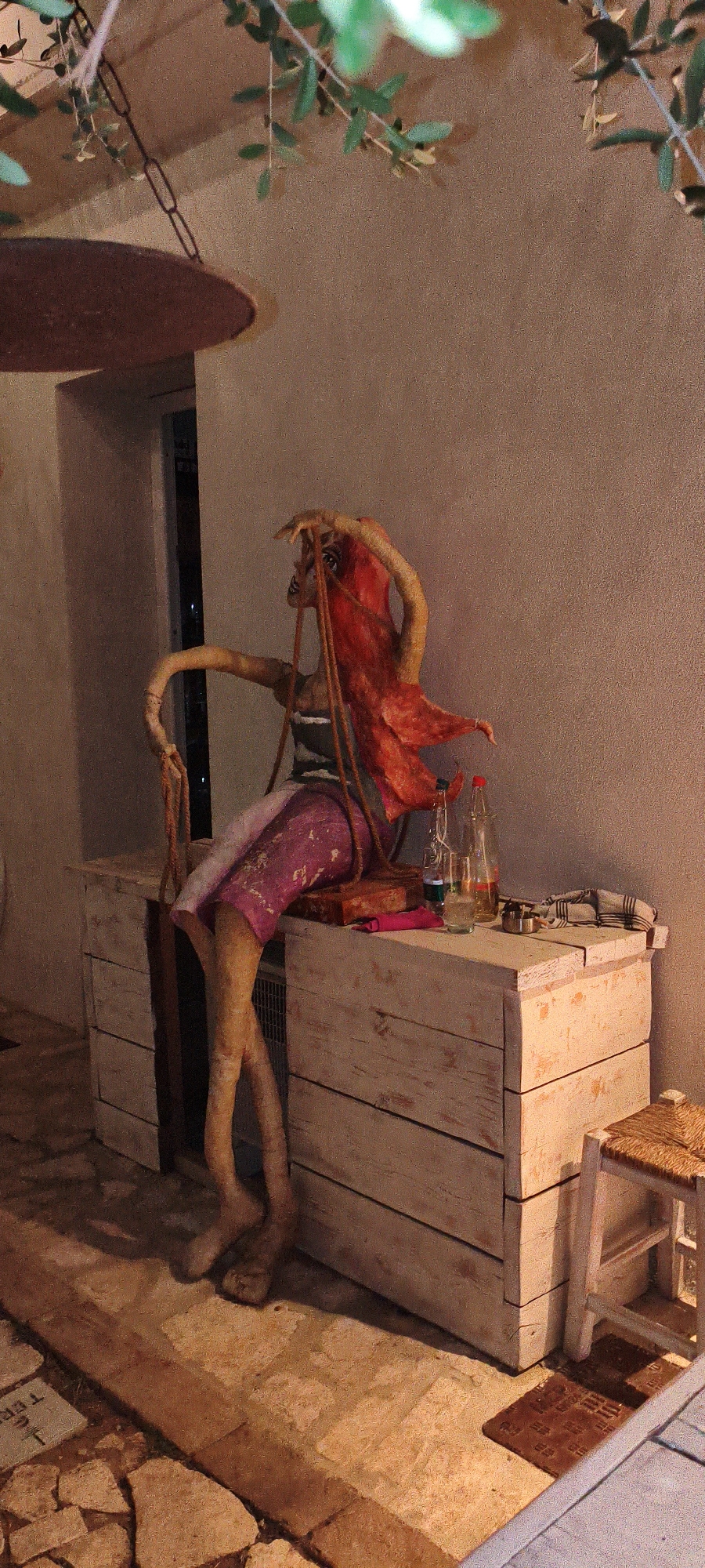 In Buriano, Italy, a peculiar mannequin-like figure, with disproportionately long, thin limbs and a vivid red headpiece resembling flowing hair, is seated on a rustic, weathered wooden chest. The figure's limbs are a pale, skin-like tone, with visible joints suggesting articulation. It wears a striped top and a pink skirt.
The wooden chest, appearing to be two stacked drawers, is painted white with visible wood grain and imperfections. On top of the chest, several objects are placed: a glass containing a clear liquid, two glass bottles with red and yellow caps, and a small ashtray. A red cloth item and a rectangular object, possibly a book or a box, are also on the chest. To the right of the chest, a small stool with a woven straw seat and white legs is positioned.
The scene is illuminated by artificial light, casting shadows. Overhead, branches with green leaves hang, some supported by a dark chain connected to a large, dark, circular object. The background is a textured, light-colored wall. The floor is paved with stones and grout, with some smaller stone debris. The overall impression is of an outdoor setting, possibly a courtyard or patio, at dusk or night.