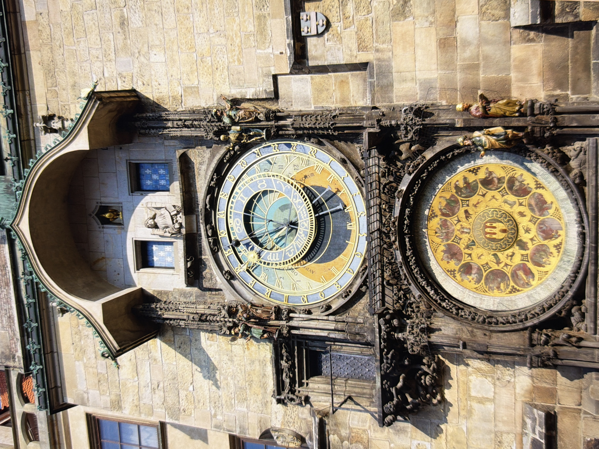 The image shows a close-up, low-angle view of the Astronomical Clock in Prague, Czech Republic. The clock tower, made of sandstone blocks, dominates the frame. The clock itself is a complex astronomical timepiece with a central dial featuring Roman numerals and intricate golden astrological symbols. Below the main clock face is a circular calendar dial adorned with round miniatures depicting scenes and zodiac signs. Two animated figures, appearing to be religious or historical characters, are positioned on either side of the main clock face. Sculptural elements and architectural details, including gargoyles and decorative stonework, are visible around the clock and on the tower. The scene is bathed in sunlight, casting distinct shadows.