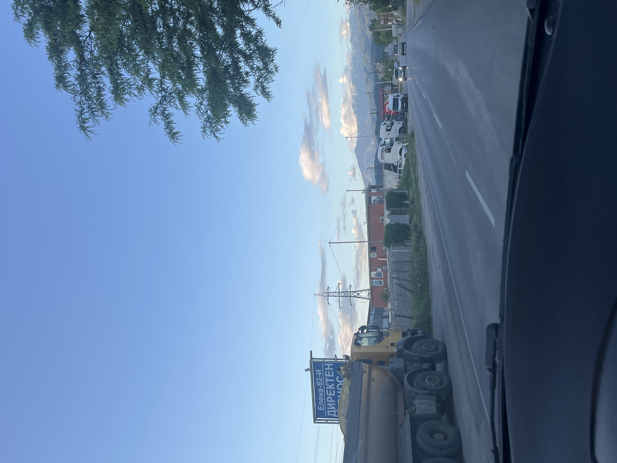 The image shows a view from inside a vehicle, possibly a car or truck, driving on a road near Blagoevgrad, Bulgaria. On the roadside, there is a large truck stopped with a sign that reads "Елена-92-и ДИРЕКТЕН ДОС" which means "Elena-92-i DIRECT DELIVERY" in Bulgarian. Behind the truck is a building and electrical transmission towers. Further down the road, there are other parked trucks and some buildings. In the background, there are distant mountains. The sky is blue with some clouds. Tree branches are visible at the top left corner of the image. The inside of the vehicle is visible on the right side of the image.