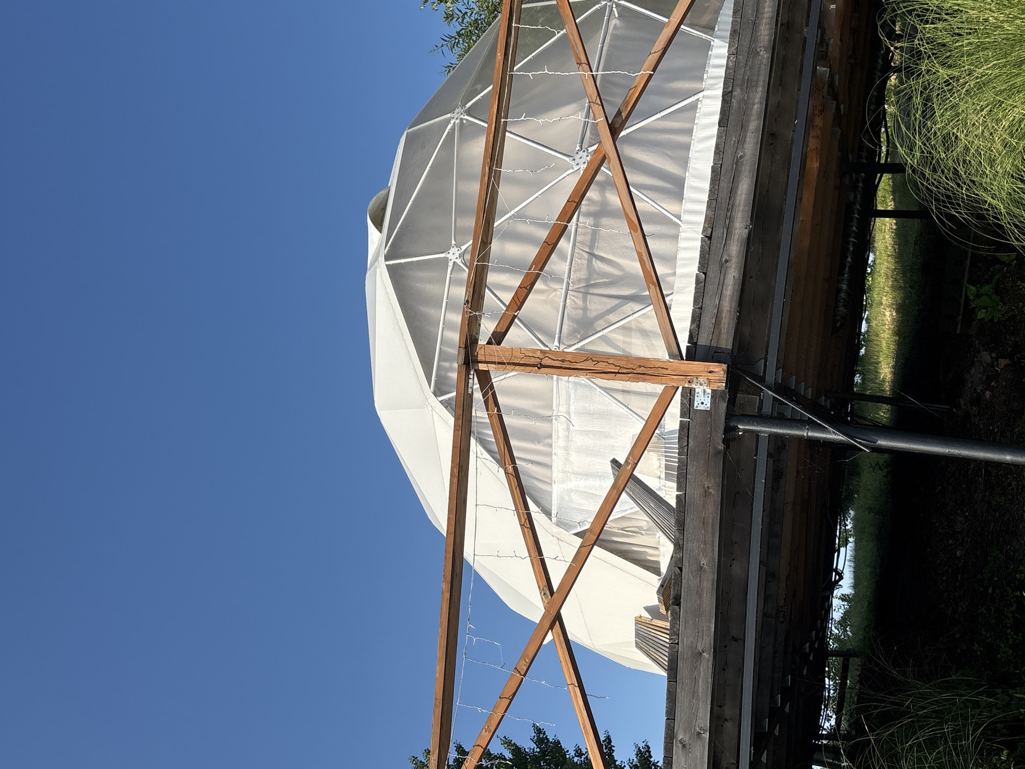 The image shows a geodesic dome structure with a translucent outer layer, partially obscured by a wooden deck in the foreground. The deck has a wooden railing with string lights attached. The geodesic dome is white and appears to be made of panels connected by a framework. The sky above is a clear blue. The setting appears to be outdoors, possibly in a rural or recreational area, given the presence of tall grass and trees. The image does not provide information about the city of Раздольное, Russia.