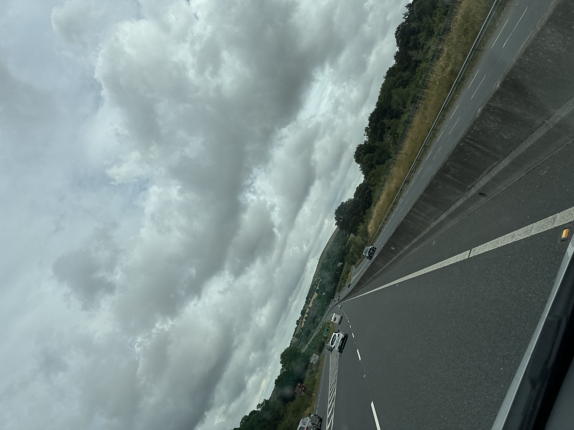 The image shows a view from a vehicle traveling on a highway in the New Ross Municipal District, Ireland. The highway has multiple lanes separated by white lines, and a concrete barrier runs along the right side. Several cars and a caravan are visible on the road. The surrounding environment consists of rolling green hills covered in trees and grass. The sky is overcast with dramatic, heavy clouds. The overall impression is a cloudy day on a scenic highway.