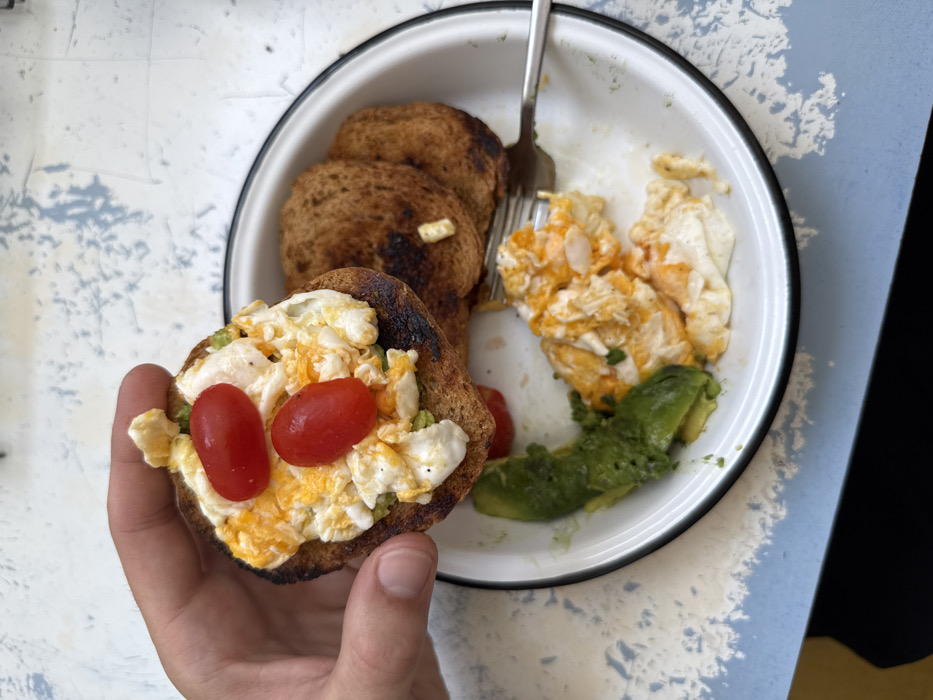 A plate of breakfast food includes toast, scrambled eggs, and mashed avocado. Two slices of toast are stacked on the plate. A third slice, topped with scrambled eggs, avocado, and two tomato halves, is held in a hand. A fork is placed on the plate next to the toast. The plate is white with a black rim. The background features a mottled surface.