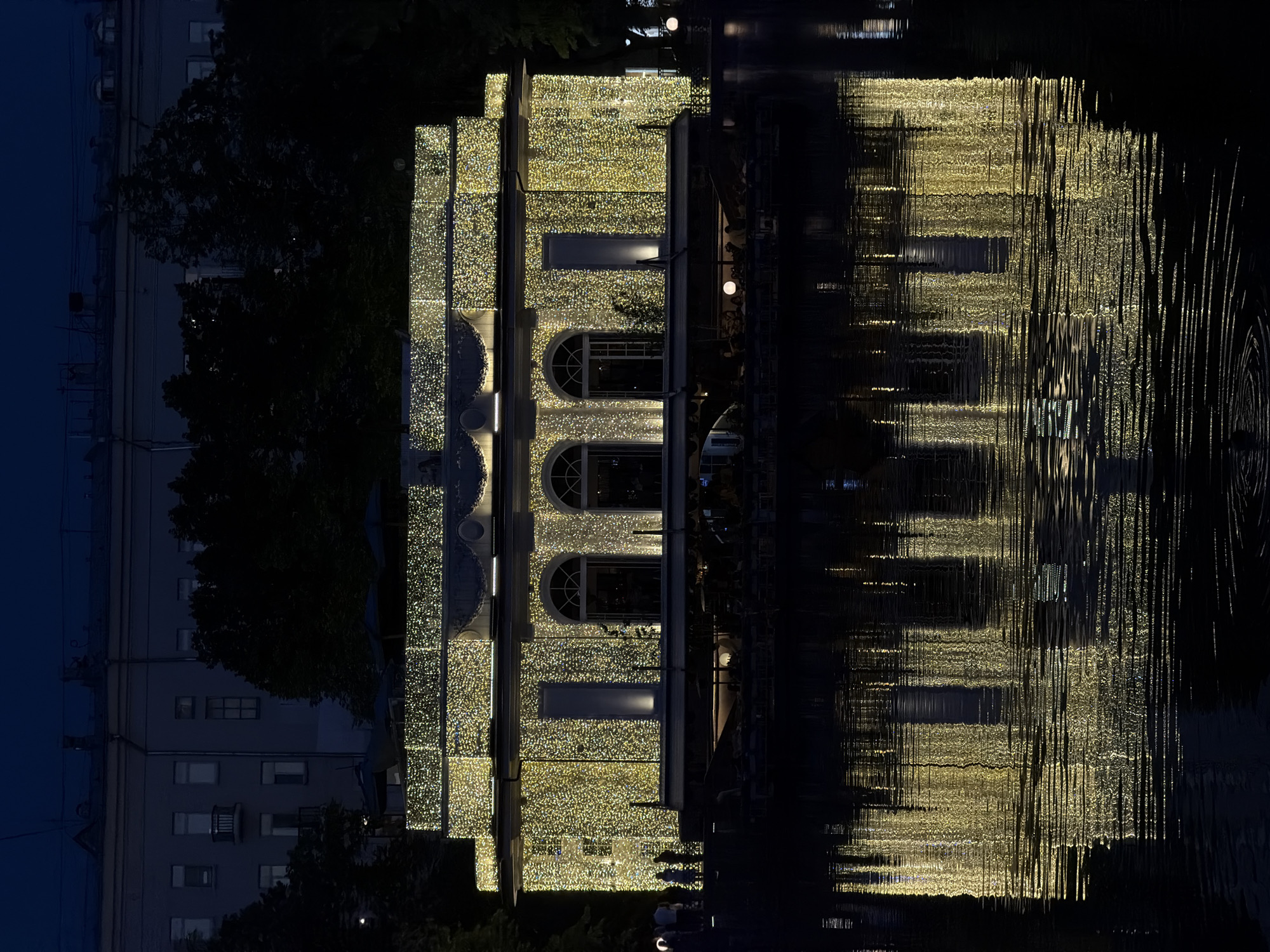 The image depicts a building illuminated with many lights, likely in Moscow, Russia, at night. The building, with its arched windows, is reflected in the water, creating a distorted mirror image. The dark sky contrasts with the bright, festive illumination of the building and its reflection. The reflection in the water is fractured and elongated due to ripples or currents, adding a textural element to the scene. A dark building stands in the background, to the left.