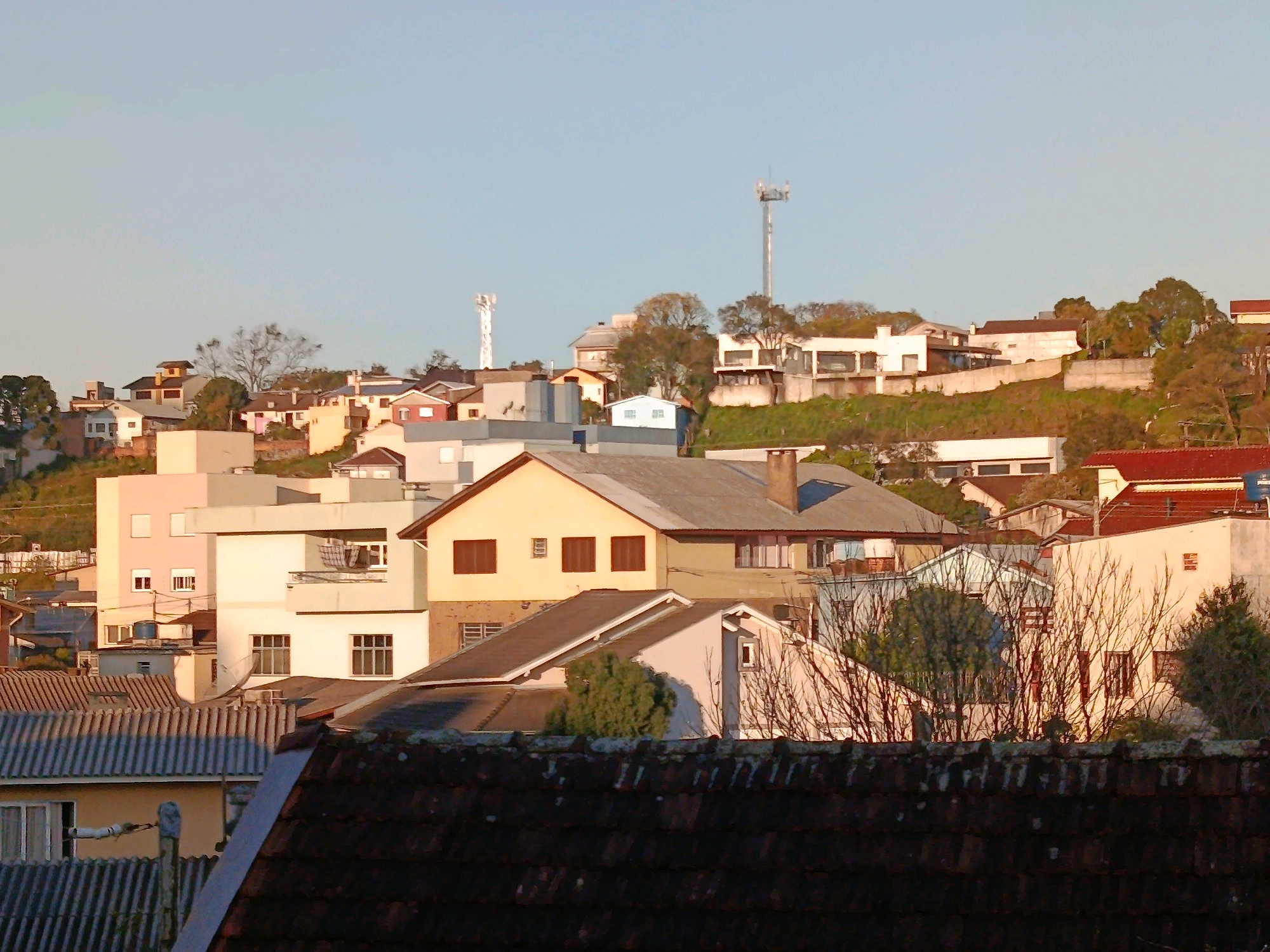 This is a landscape photograph of a residential area in Caxias do Sul, Brazil, taken on a clear, sunny day. The image captures a dense collection of houses and buildings nestled on a hillside, with the sky a pale, clear blue. The warm, golden light of what appears to be late afternoon or early morning illuminates the scene, casting soft shadows. In the foreground, a dark-tiled roof occupies the lower portion of the frame. Beyond this, a jumble of various residential structures with different roof types, including corrugated metal and tile, are visible. Several houses have multiple stories, and architectural styles vary, with some featuring balconies and prominent windows. Two prominent communication towers are visible in the background, one taller and more central than the other, situated on the crest of the hill. The hillside is covered with vegetation, including trees and shrubs, some of which are bare, suggesting a cooler season. The overall impression is of a moderately developed urban or suburban neighborhood on uneven terrain.