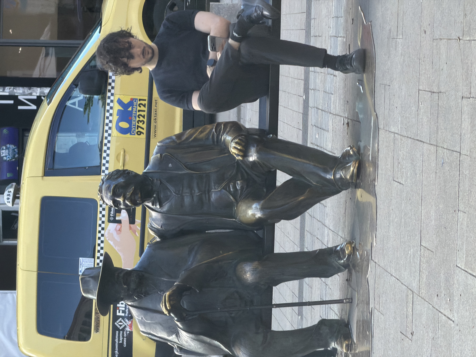 The image captures a vibrant urban street scene in Sofia, Bulgaria, during bright daylight. It features a young man seated on a stone bench alongside two bronze statues, with a yellow taxi positioned behind them.
The man, with dark, curly hair and a beard, is wearing a black t-shirt, dark pants, black socks, and dark shoes. He is sitting casually on the bench, engrossed in his smartphone, which he holds in both hands. A smartwatch is visible on his left wrist.
To his right, two life-sized bronze statues of men are also seated on the bench. The statue closer to the foreground is depicted wearing a hat and holding a cane. Both statues exhibit the characteristic green and brown patina of aged bronze, with certain areas, particularly on their hands, knees, and feet, appearing polished and brighter, suggesting frequent touching by passersby.
Behind the bench, a bright yellow taxi with a distinctive black and white checkered stripe on its side is visible. The taxi displays several pieces of text: "OK СУПЕРТРАНС" (OK Supertrans), a phone number "9732121", and a website "www.oktaxi.net". Towards the rear of the taxi, an advertisement for "Fibar" shows a hand holding a card, accompanied by the Bulgarian text "Безконтактни карти само от..." (Contactless cards only from...). The number "1935" is also discernible near the back window. In the background, partially obscured, is a blue object with the "Monster" energy drink logo.
The ground is paved with light grey, rectangular tiles, indicating a well-maintained public space. The scene conveys a casual, everyday moment in a bustling city.