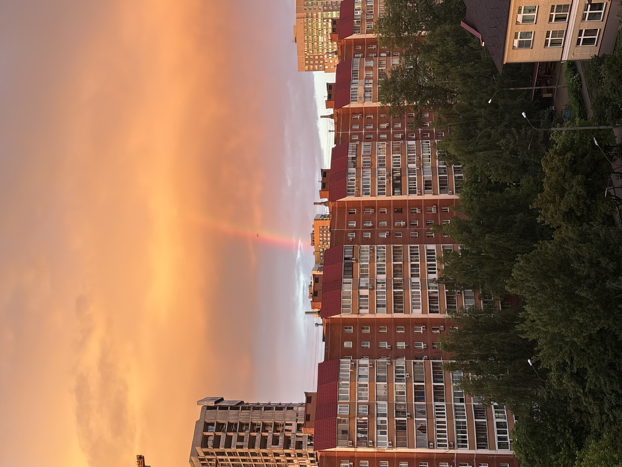 This is an outdoor, eye-level, medium shot of a city skyline in Mytishchi, Russia, during a sunset. The sky is filled with vibrant orange and pink clouds, creating a dramatic backdrop. A faint rainbow is visible in the center of the frame. In the foreground, several multi-story brick apartment buildings with red roofs dominate the scene. A partially constructed concrete building stands to the left, and a taller, modern apartment building is visible on the far right. Dense green trees partially obscure the lower portions of the apartment buildings, and streetlights are visible throughout the scene. No people are clearly visible in the image. The overall impression is one of a residential urban landscape bathed in the warm, dramatic light of a sunset.