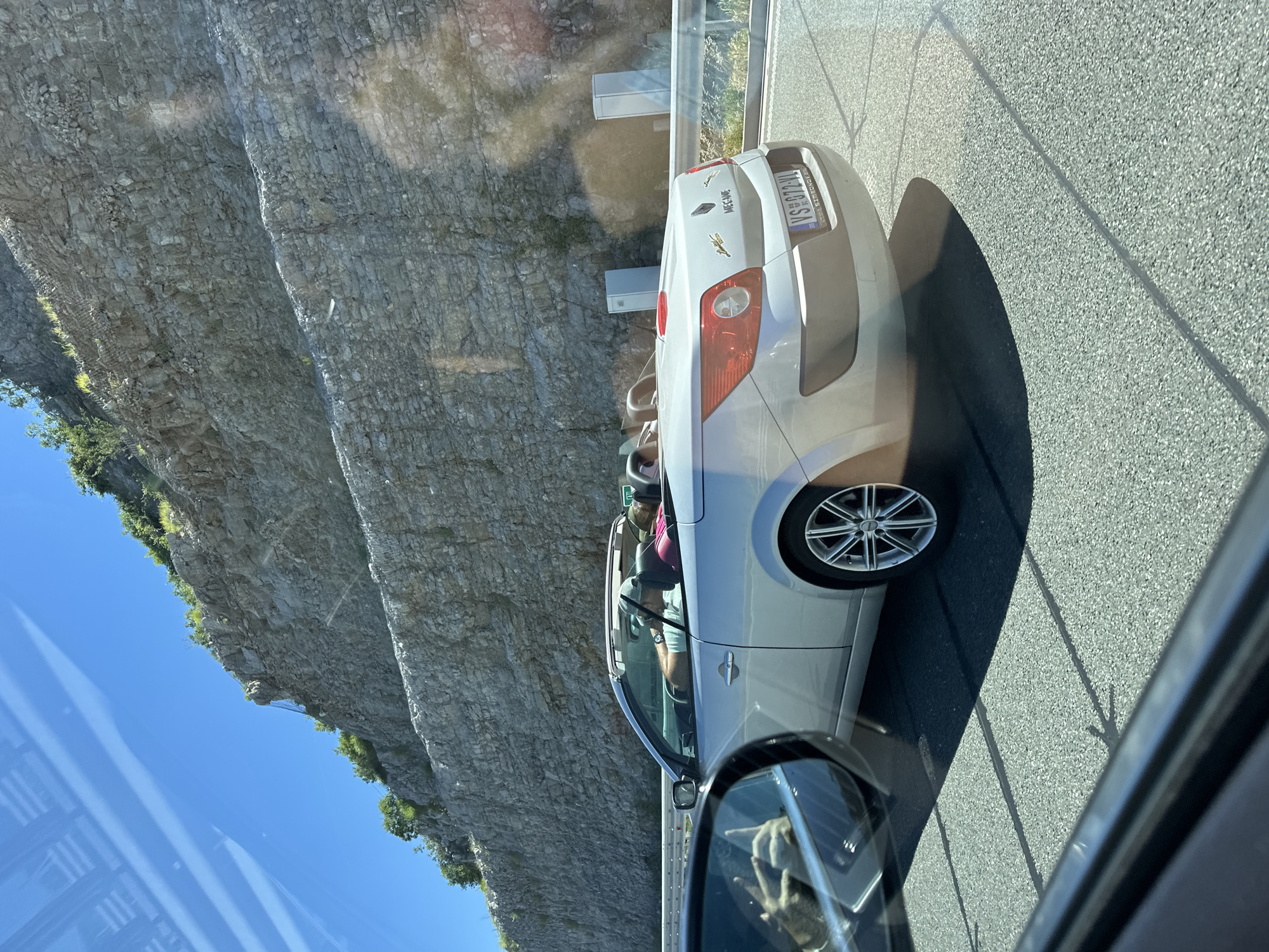 A silver Renault Megane convertible is driving on a highway in Klopot, Montenegro. The car has its top down, and two people are visible inside. A man is driving, wearing a light-colored t-shirt and a baseball cap, and a person with pink hair is in the passenger seat, wearing a dark top. The car's license plate reads "VS 872 VL". The highway is flanked by a steep, rocky cliff face on the right and a metal guardrail. In the foreground, the driver's side mirror of another vehicle reflects the hand of the photographer, which is giving a peace sign. The road surface is asphalt with visible lane markings. The sky is clear and blue, and the sun is casting shadows.