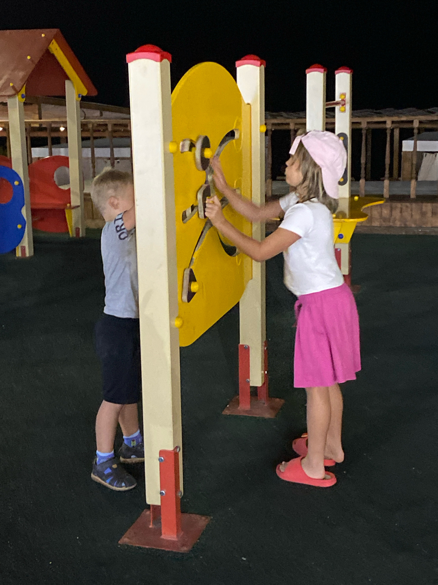 Two children are interacting with a yellow playground activity panel. A boy, wearing a light blue t-shirt and dark shorts, stands to the left of the panel, his body partially obscured. A girl, wearing a white t-shirt and a bright pink skirt, stands to the right, her hands engaged with the panel. She is also wearing a pink hat. The playground equipment is set against a dark background, suggesting it is nighttime. The ground surface is black. In the background, other playground structures are visible, including a red and yellow playhouse. The overall scene appears to be an outdoor playground.