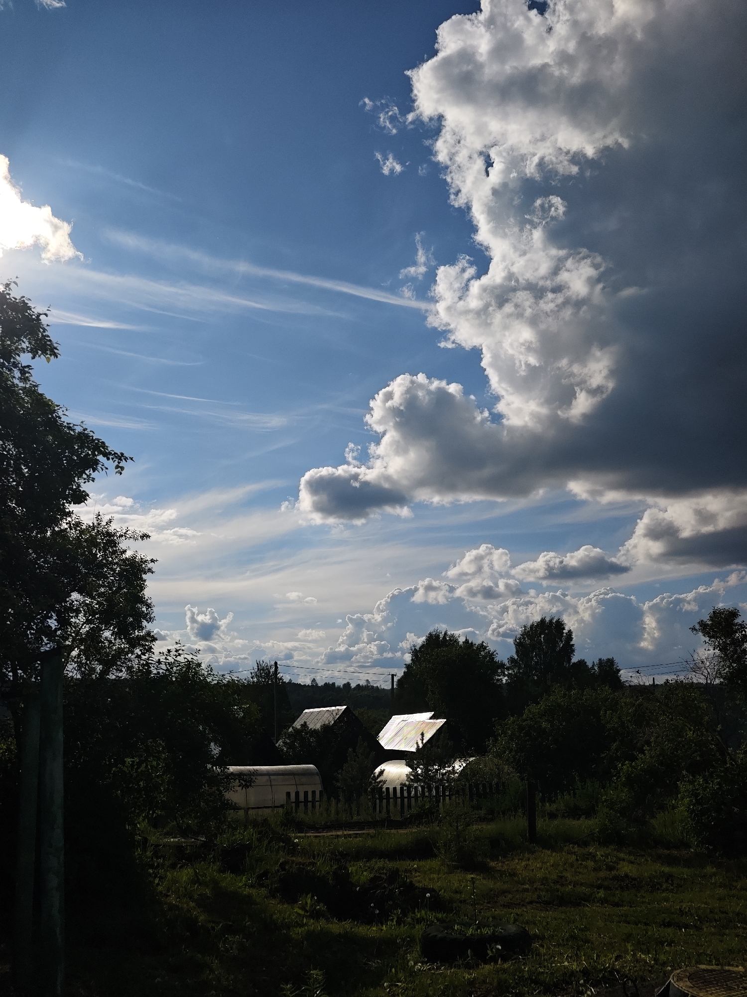 The image depicts a rural landscape in Уральское, Russia. The sky dominates the scene, featuring a mix of bright blue patches and dynamic cloud formations, including cumulus and cirrus clouds. Some of the clouds are dark, suggesting an approaching storm.
In the lower part of the frame, there is a view of a backyard or garden. There are several buildings with metallic roofs, partially obscured by trees and dense foliage. The foliage is dark, indicating possible shadows. The ground is covered in grass and possibly weeds, typical of a rural setting. The light suggests it is daytime.